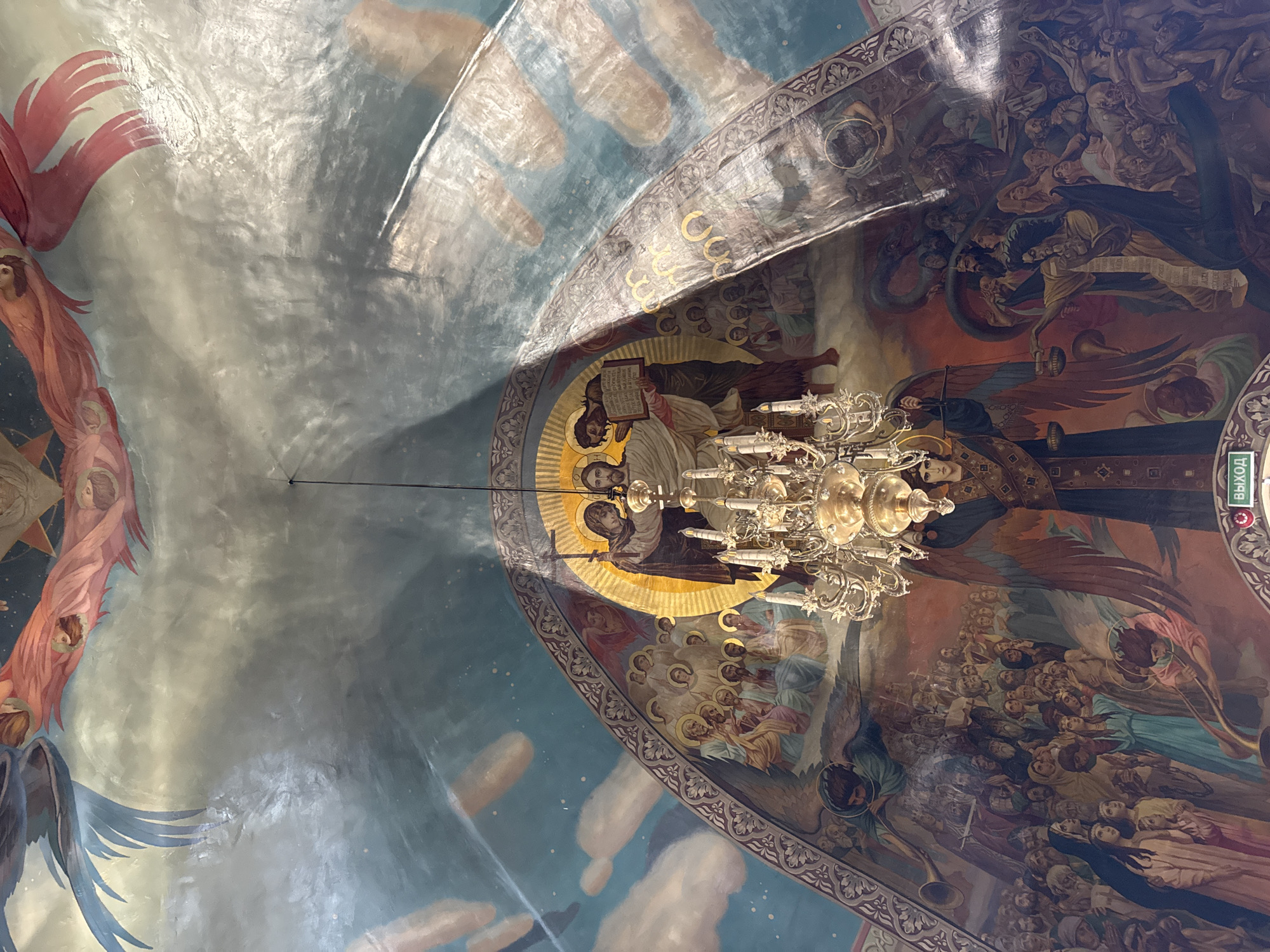 This is a low-angle, full shot of a frescoed ceiling and upper walls of a church or cathedral. The artwork depicts religious scenes with multiple figures. In the center, a large, ornate chandelier hangs from the ceiling. The fresco depicts a central scene of Jesus and another figure, likely John the Baptist, surrounded by saints and angels. Below this, the artwork continues to depict a large gathering of people, some with halos, possibly representing the resurrection or judgment day, with angels blowing trumpets. On the right side of the fresco, a more chaotic scene unfolds with figures being pulled into a swirling abyss, a large serpent, and figures with demonic features. The artwork is rendered in vibrant colors and detailed depictions of human forms and drapery. The environment appears to be an interior space with a high, vaulted ceiling, decorated with painted clouds and what appear to be disembodied hands in the upper portions of the fresco. There is a sign in Russian text at the bottom of the frame.