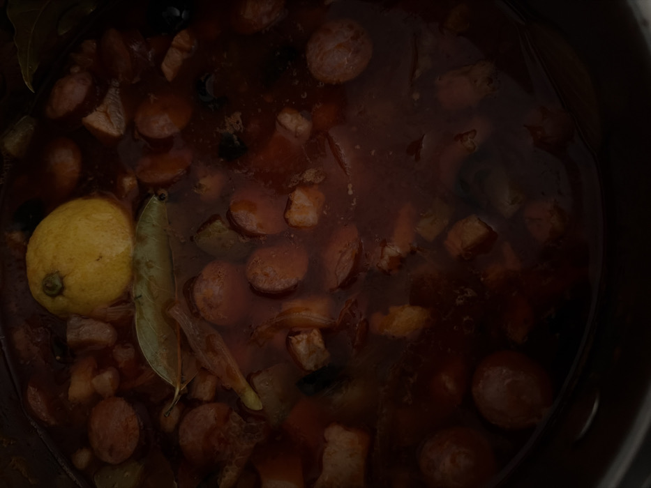 An overhead view depicts the contents of a dark cooking pot, located in Novosaratovka, Russia. The pot contains a dark reddish-brown liquid broth with various solid ingredients submerged and floating. A whole, light yellow lemon with a dark stem is positioned in the upper-central portion of the frame, partially submerged. Below the lemon, a single olive-green bay leaf floats on the liquid surface. Numerous circular slices of reddish-brown sausage are visible throughout the pot, along with irregularly shaped pieces of light brown meat. Scattered dark, oval-shaped objects, consistent with olives, are also present. The scene is dimly lit, with the dark rim of the pot visible along the edges. No individuals are present.
