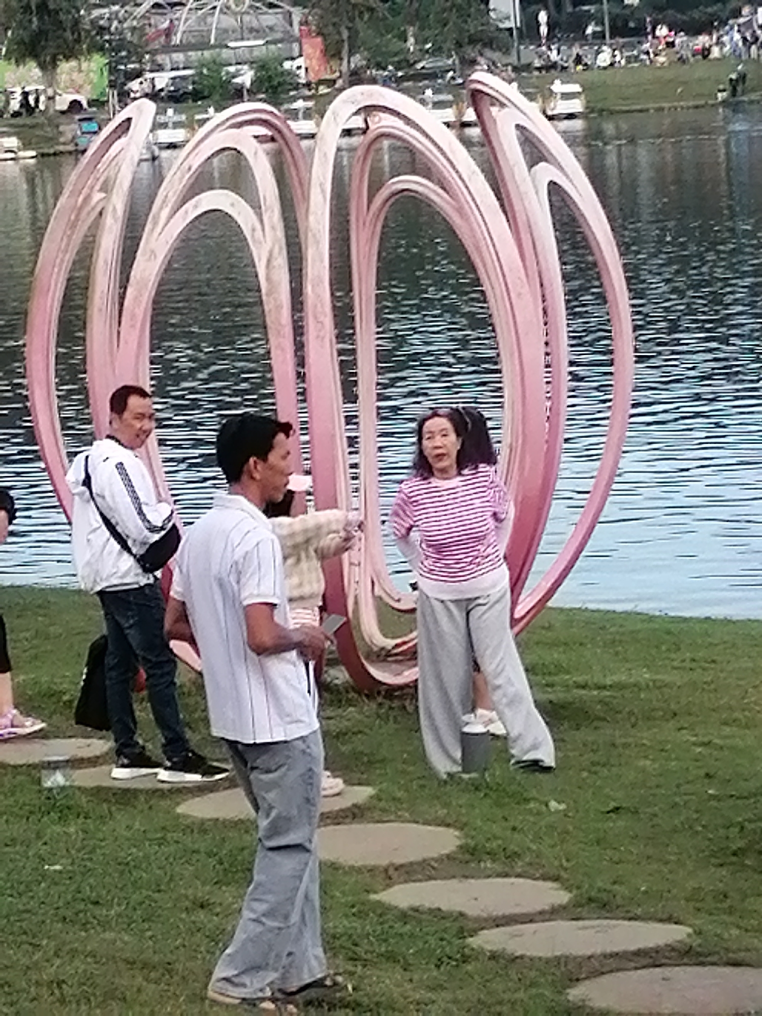 In Da Lat, Vietnam, a group of people are seen near a large, pink, abstract sculpture resembling intertwined loops, situated on a grassy area adjacent to a body of water. The sculpture is the central focus, with its organic, flowing design dominating the background. In the foreground, on the right, a woman in a pink and white striped shirt and grey pants stands with her hands on her hips, looking towards the viewer. To her left, a man in a white short-sleeved shirt with vertical stripes and light blue jeans is walking away, holding a dark object in his hand. Further to the left, another man wearing a white jacket with black trim and dark jeans is looking back and smiling. Behind him, a person is partially visible. The ground is covered in green grass, with a path of stepping stones leading away from the sculpture. The water in the background reflects the surrounding trees and the sky, creating a sense of depth. The overall scene appears to be a public outdoor space, likely a park or recreational area, where visitors are interacting and enjoying the environment.