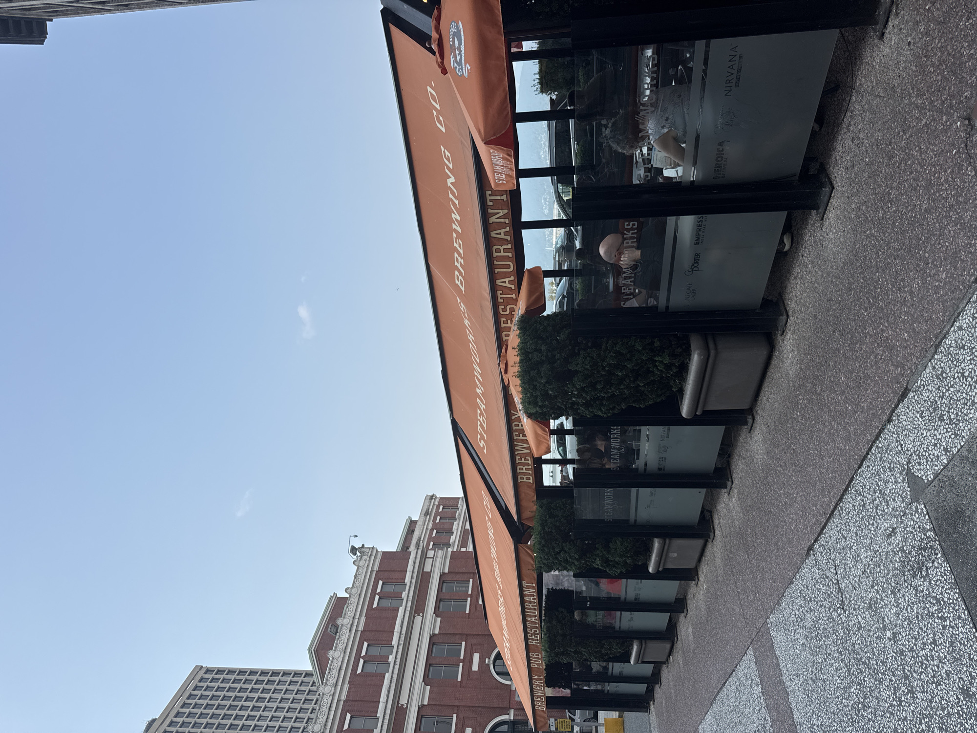 The image shows an outdoor seating area of Steamworks Brewing Co. in Vancouver, Canada. The restaurant has orange awnings with "BREWERY PUB RESTAURANT" and "STEAMWORKS BREWING CO." printed on them. There are glass barriers with frosted graphics, some of which display beer names like "EMPRESS INDIA PALE ALE" and "NIRVANA INDIA PALE ALE." Green bushes are planted in rectangular planters along the edge of the seating area. Inside the seating area, several patrons are visible, some dining and others engaged in conversation. The background features a large brick building and a modern high-rise building, suggesting an urban setting. The sidewalk in front of the restaurant is paved with a speckled material. The sky is clear and bright.