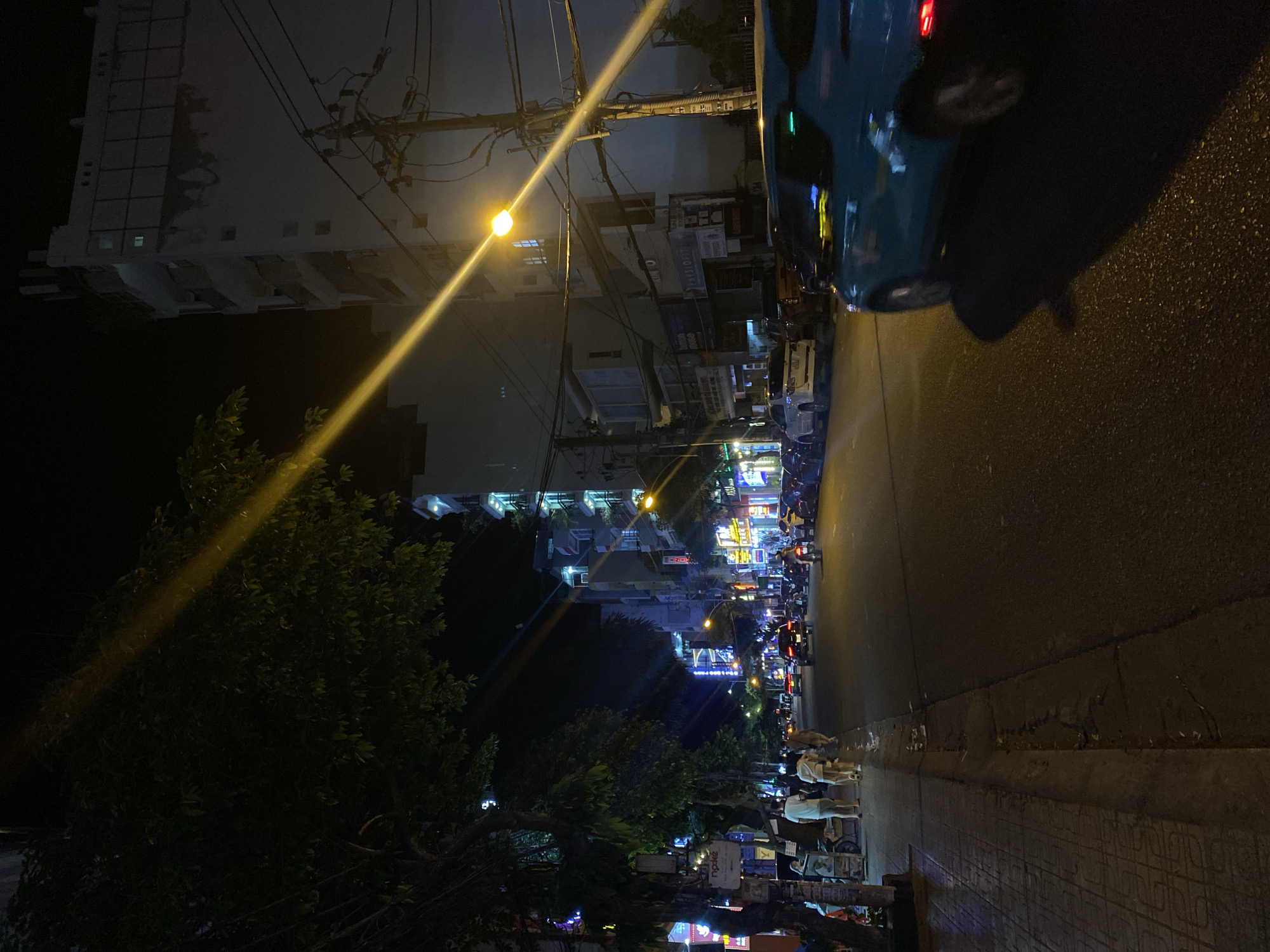 The image captures a nighttime street scene in Nha Trang Ward, Vietnam, likely a commercial or residential area. A bright streetlamp casts a strong diagonal beam across the frame, illuminating part of a multi-story building with a white facade and visible windows. In the foreground, a turquoise car is captured in motion, its rear taillights illuminated. The street is lined with parked cars and motorcycles, suggesting moderate traffic. Pedestrians, including a group of three individuals walking on the sidewalk on the left, are present. The sidewalk features a tiled pattern and is bordered by trees with dark foliage. Buildings along the street are lit with various signs, some displaying neon lights. Overhead, a complex network of electrical wires is visible. The overall impression is a vibrant, active urban environment at night, with characteristic street lighting and infrastructure.