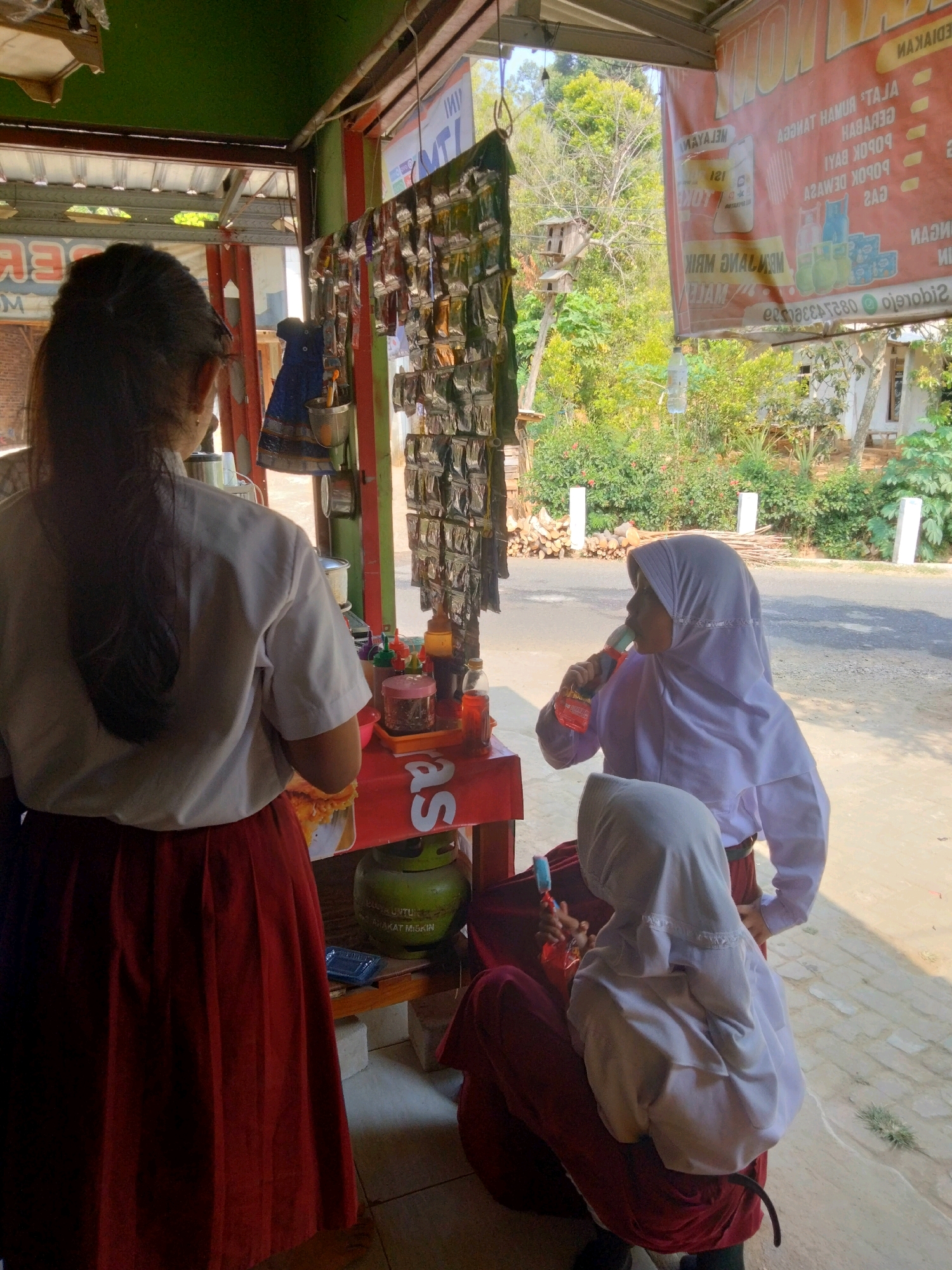 In Sidorejo, Indonesia, three girls wearing school uniforms stand at an outdoor food stall. The girl on the far left, with long dark hair, faces away from the camera, her back to the viewer. She wears a white shirt and a red skirt. To her right, a girl in a light purple hijab and matching top is drinking from a red and blue bottle. She is seated on the ground. Next to her, another girl in a white hijab and dark red skirt is also seated. The food stall has a red counter with various items on display, including small packets hanging from a rack. A green gas cylinder is visible behind the counter. In the background, there is lush greenery, trees, and a road. A large red banner with Indonesian text is hung above the stall.