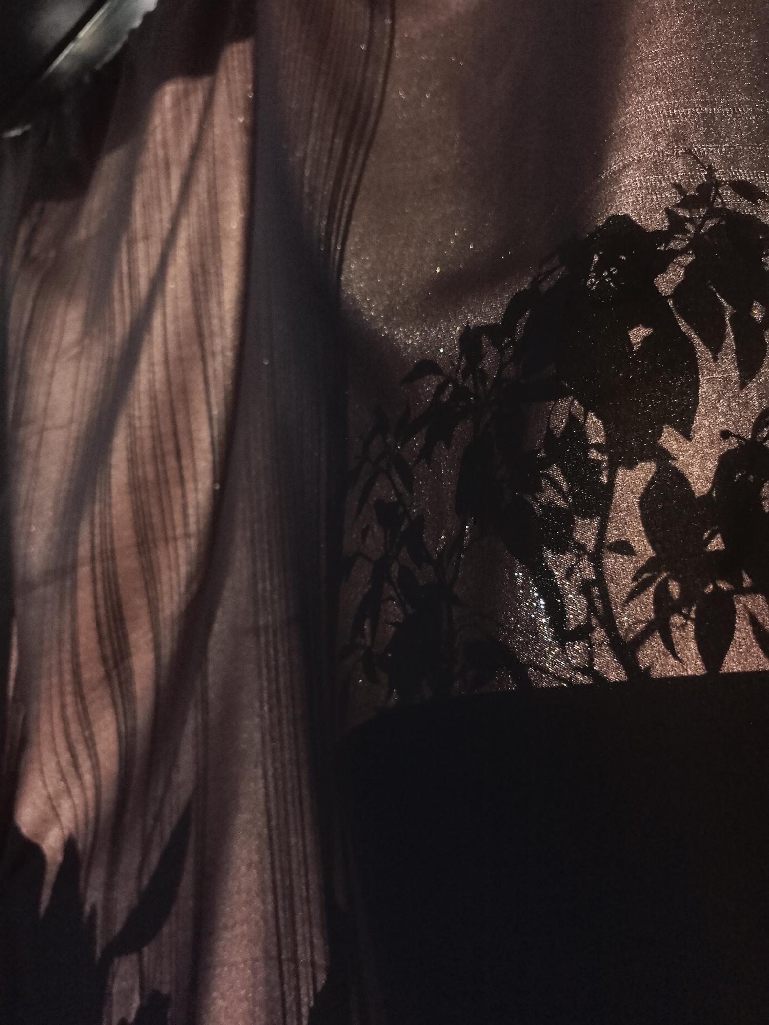 The image shows a close-up view of a sheer, textured fabric with a vertical striped pattern on the left side and a shadow cast by leaves or foliage on the right. The fabric appears to be draped or hanging. The overall tone of the image is dark, with the fabric being illuminated by a subtle light source. No people or specific objects are discernible beyond the fabric and shadow. The location is not evident within the image, and the provided city name is unlikely to be relevant.