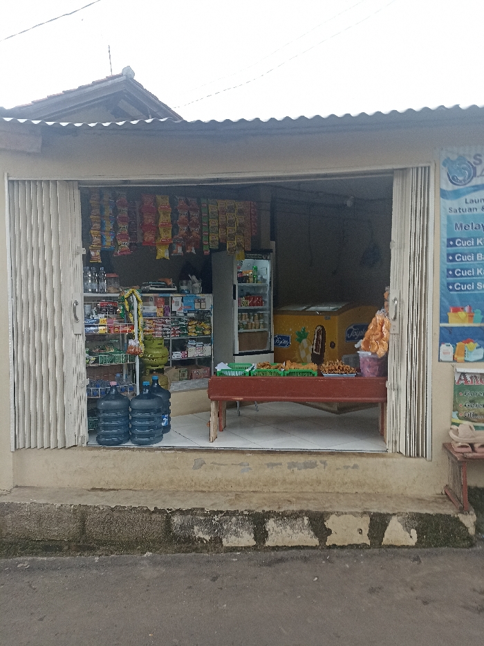 The image shows the front of a small store in Jatinangor, Indonesia (latitude: -6.936, longitude: 107.779). The store has a white corrugated metal roof and white painted walls. The entrance is open, with a wooden bench and a table, both of which are covered in plastic crates. The table is stacked with various snacks and goods. To the left of the entrance, two blue water jugs sit in front of a metal shelf holding many packaged goods. On the right side of the entrance, a sign is mounted on the wall listing the services offered at the store: Laundry, Unit & Laundry, and several options for laundry services. The sign is written in Indonesian and reads: "Laundry Satuan & Kiloan, Melayani: Cuci Kiloan, Cuci Bed Cover, Cuci Kursi, Cuci Seprei." The store is located in a residential area, with a paved road and a walkway in front of the store.