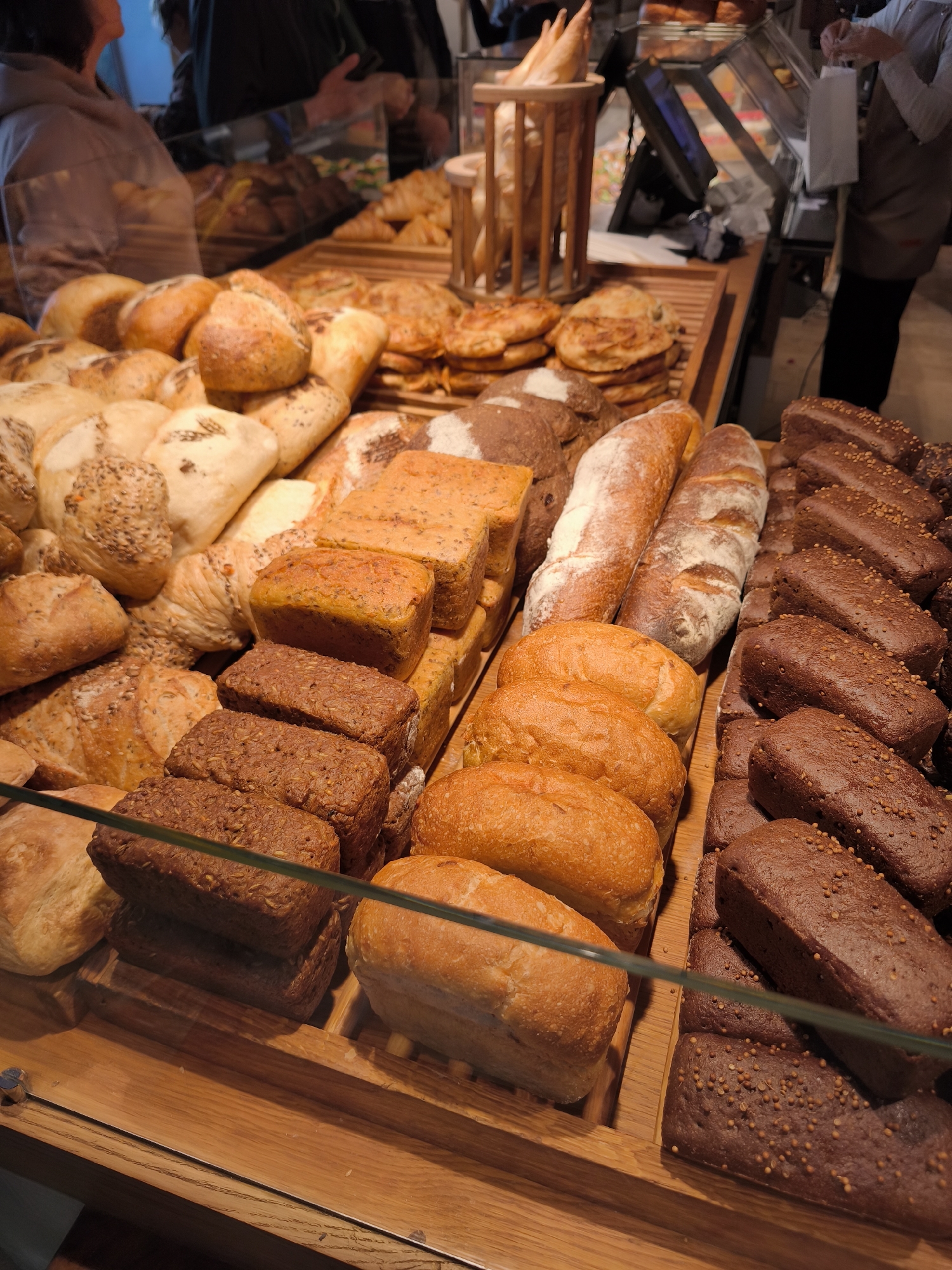 The image shows a display of various types of bread in a bakery. The bread includes loaves, rolls, and baguettes, with different textures and toppings. Some loaves are light in color, while others are dark brown. Several loaves have visible seeds or grains on their crusts. The bread is arranged on wooden shelves behind a glass counter. In the background, a person is visible, partially obscured, reaching towards a display case. The environment appears to be a food establishment, likely a bakery or cafe. The scene is brightly lit, highlighting the textures and colors of the bread. The specific location of Svetlogorsk, Russia, is not directly indicated by any visual cues in the image.