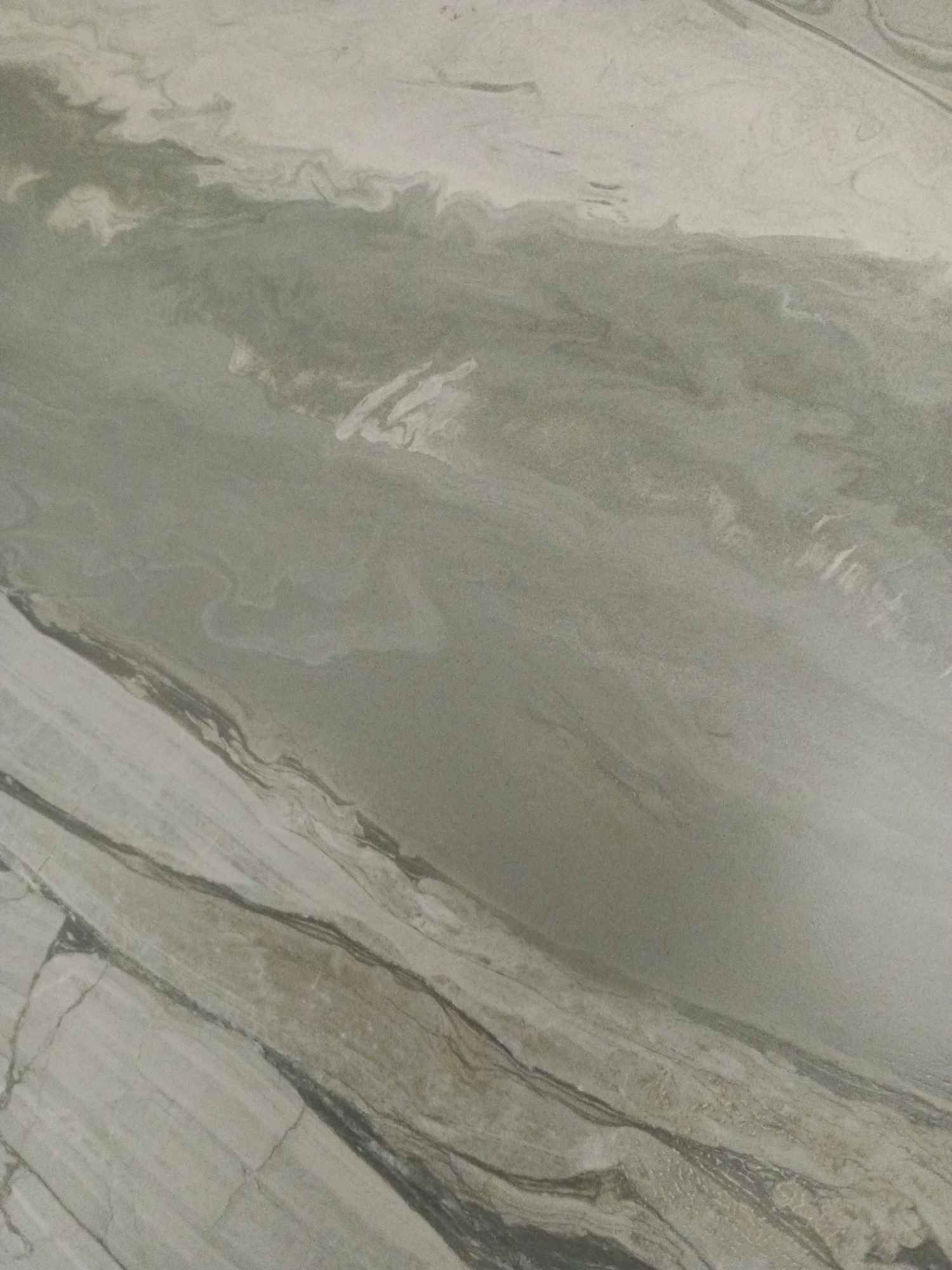 The image is a close-up, high-angle shot of a textured surface, likely marble or a similar stone. The surface exhibits a pattern of swirling veins and irregular lines in varying shades of grey, white, and muted green. The texture appears to be matte rather than polished. There are no people, objects, or discernible environmental features present in the image, making it impossible to determine its location within the city of Biysk, Russia, or any other specific place. The image exclusively depicts the abstract patterns of the stone material.