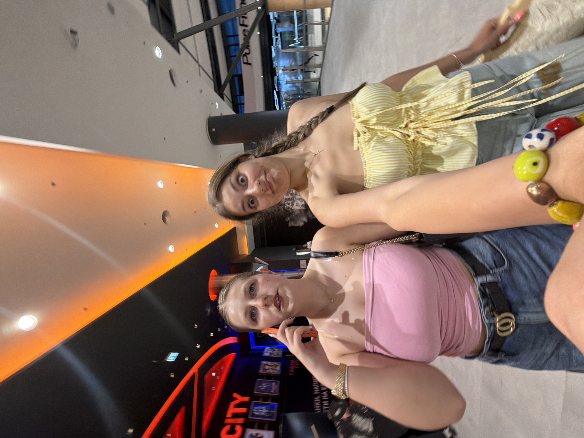 Two young women are depicted indoors, likely within a modern commercial or entertainment complex in Burgas, Bulgaria. The image is taken at a slight upward angle, with the subjects filling much of the frame.
The woman in the foreground, on the left side of the image, has blonde hair and blue eyes. She is wearing a light pink strapless top and denim shorts or a skirt with a black belt featuring a gold buckle. She has gold bracelets on her right wrist and is holding an orange smartphone to her left ear, appearing to be engaged in a phone conversation. Her expression is attentive.
Behind and slightly to the right of her is another young woman with long brown hair styled in a braid over her left shoulder. She is wearing a yellow and white striped strapless top with decorative ties. Her lower body is clad in denim jeans. She is making a playful or funny face directly at the camera, with wide eyes and pursed lips. A prominent, colorful beaded bracelet is visible on her left wrist.
The setting features minimalist architecture with light grey/white walls and dark accent walls with orange illuminated strips. Recessed lighting fixtures are visible on the ceiling. In the background, behind the woman in yellow, part of a sign with "PureFi" is visible. On the dark wall behind the woman in pink, large red letters spell out "CITY". Below this, more text is visible, including partially obscured Cyrillic script such as "АНКИ, НАРЮ" and "НА НАР", suggesting an entertainment venue like a cinema or mall directory. Several framed images or posters are displayed on a vertical panel. The floor is light-colored. The time of day is not discernible due to the indoor lighting.