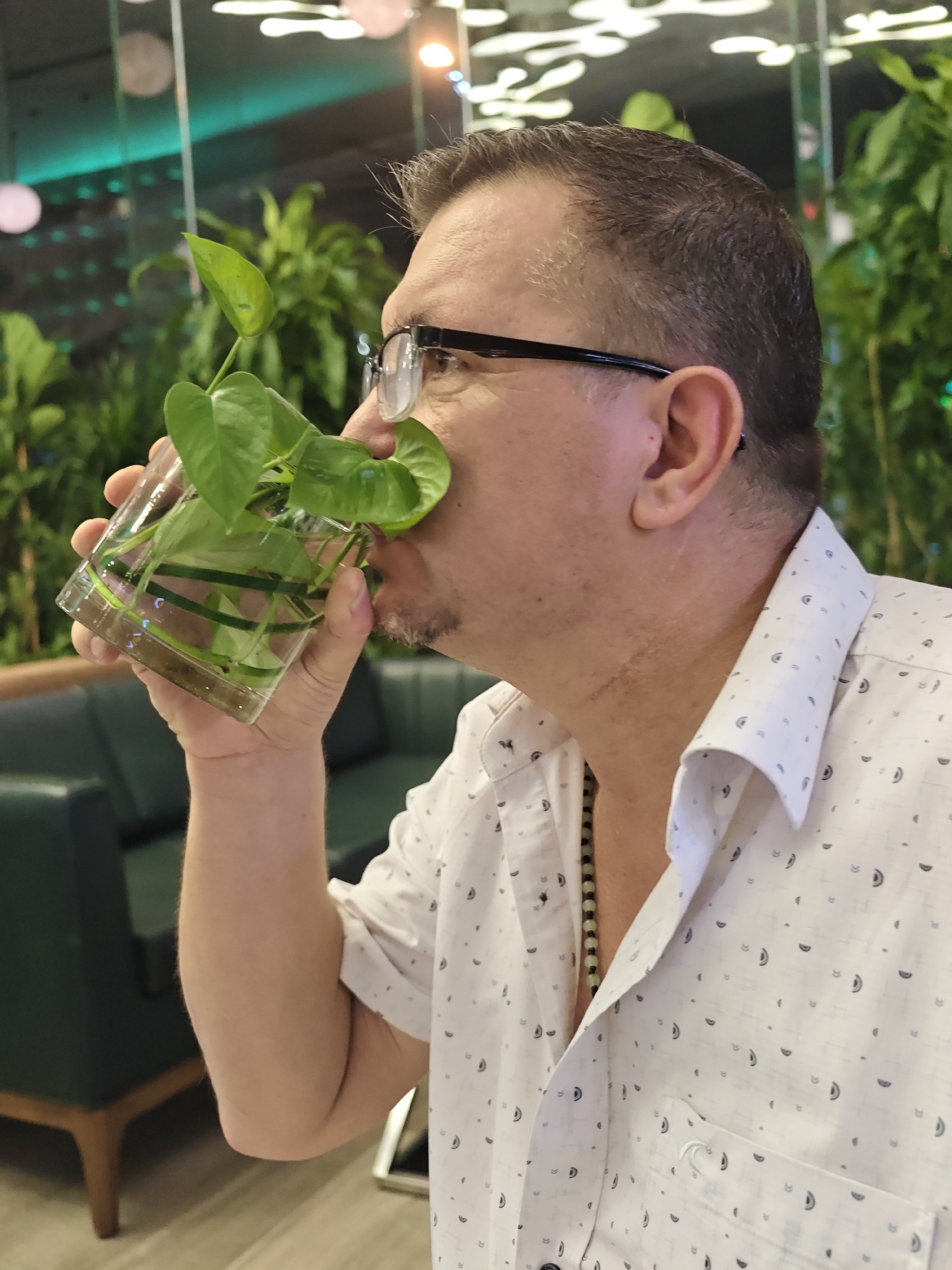 A medium shot captures a man in profile, holding a clear glass of water with a small green plant rooted in it, close to his face. He is wearing a white collared shirt with a subtle pattern of dark crescent shapes and small dots. A beaded necklace is visible around his neck. His short, dark hair is thinning on top, and he wears dark-rimmed glasses. The man has a light beard and mustache.
The background suggests an indoor setting, possibly a restaurant or cafe, with lush green plants visible behind him. Soft, ambient lighting illuminates the scene, with some out-of-focus circular lights appearing in the upper portion of the image. A dark green sofa is partially visible in the mid-ground to the left. The overall impression is a casual moment captured during the day, with no specific indicators of time or weather. The man's expression is neutral as he appears to be smelling or examining the plant. The location context of Nha Trang Ward, Vietnam, is provided externally.