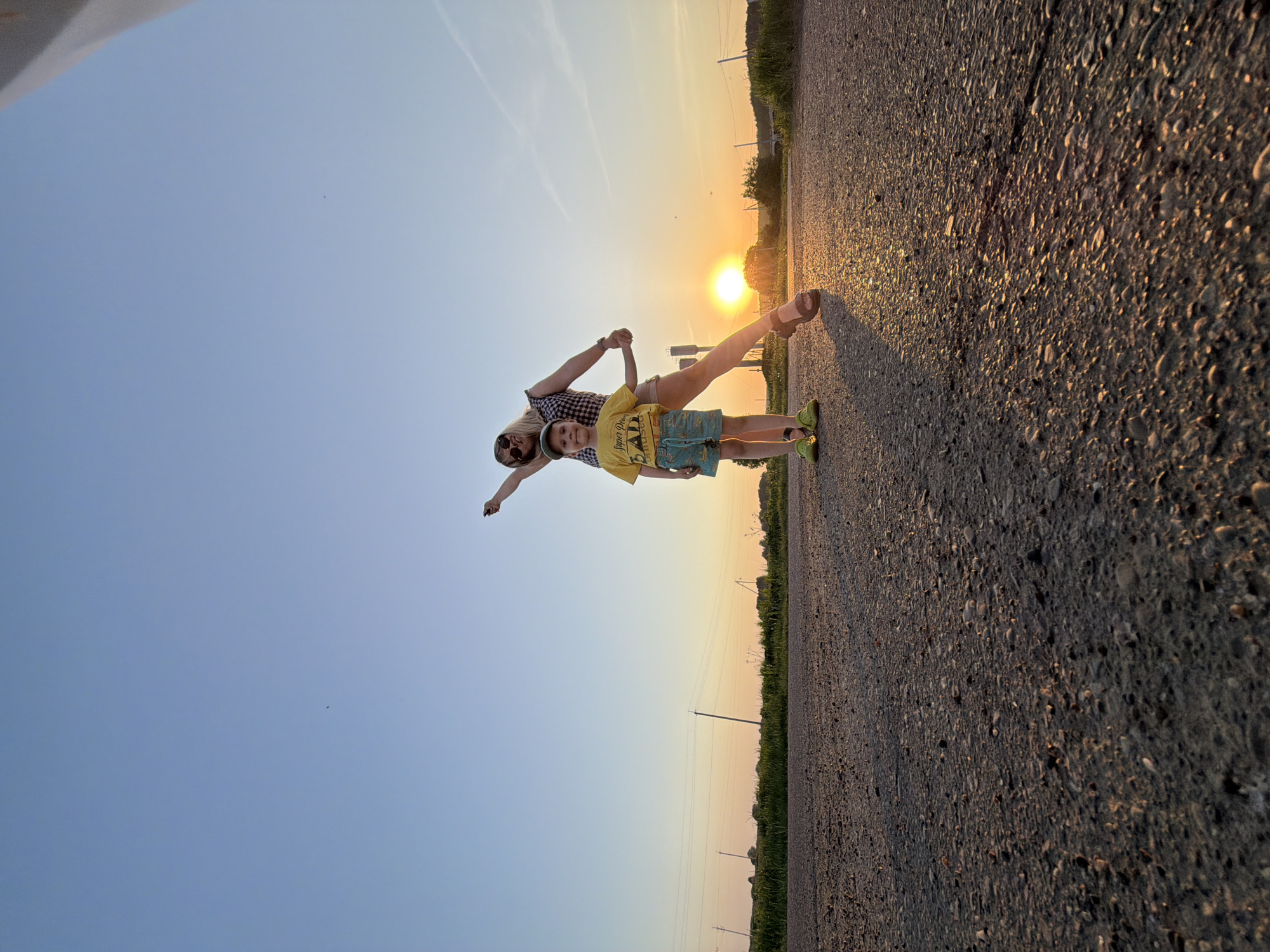 A young boy and an adult are standing on an asphalt road surface during what appears to be sunset or sunrise. The boy is wearing a yellow t-shirt and blue shorts. The adult is wearing a patterned shirt and shorts, and they are holding hands while facing the sun. Both have their arms outstretched, mimicking an airplane. The location, according to the prompt, is in Чумна, Russia. The surrounding environment includes fields and power lines visible in the background, along the edge of the road. The sky is clear with a gradient of color from blue to orange near the horizon.