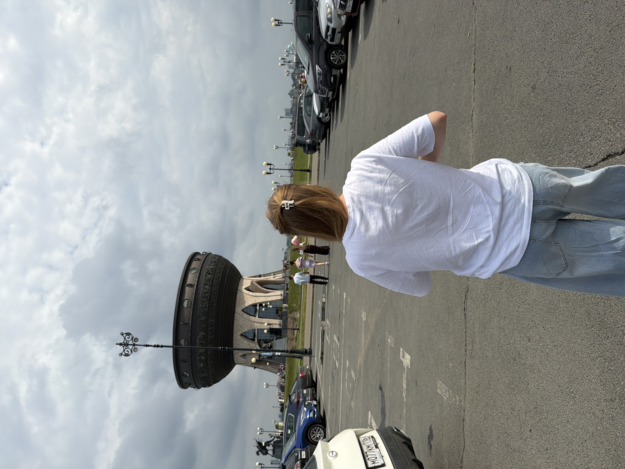 A woman with blonde hair pulled back with a decorative clip stands in a parking lot facing away from the camera. She is wearing a white t-shirt and light blue wide-leg jeans. In the background, a large, distinctive building with a wide, cauldron-like structure at the top stands prominently. This building is the Palace of Farmers, a well-known landmark in Kazan, Russia. Several cars are parked in the lot, and a few people can be seen walking in the distance near the building. The sky is cloudy.