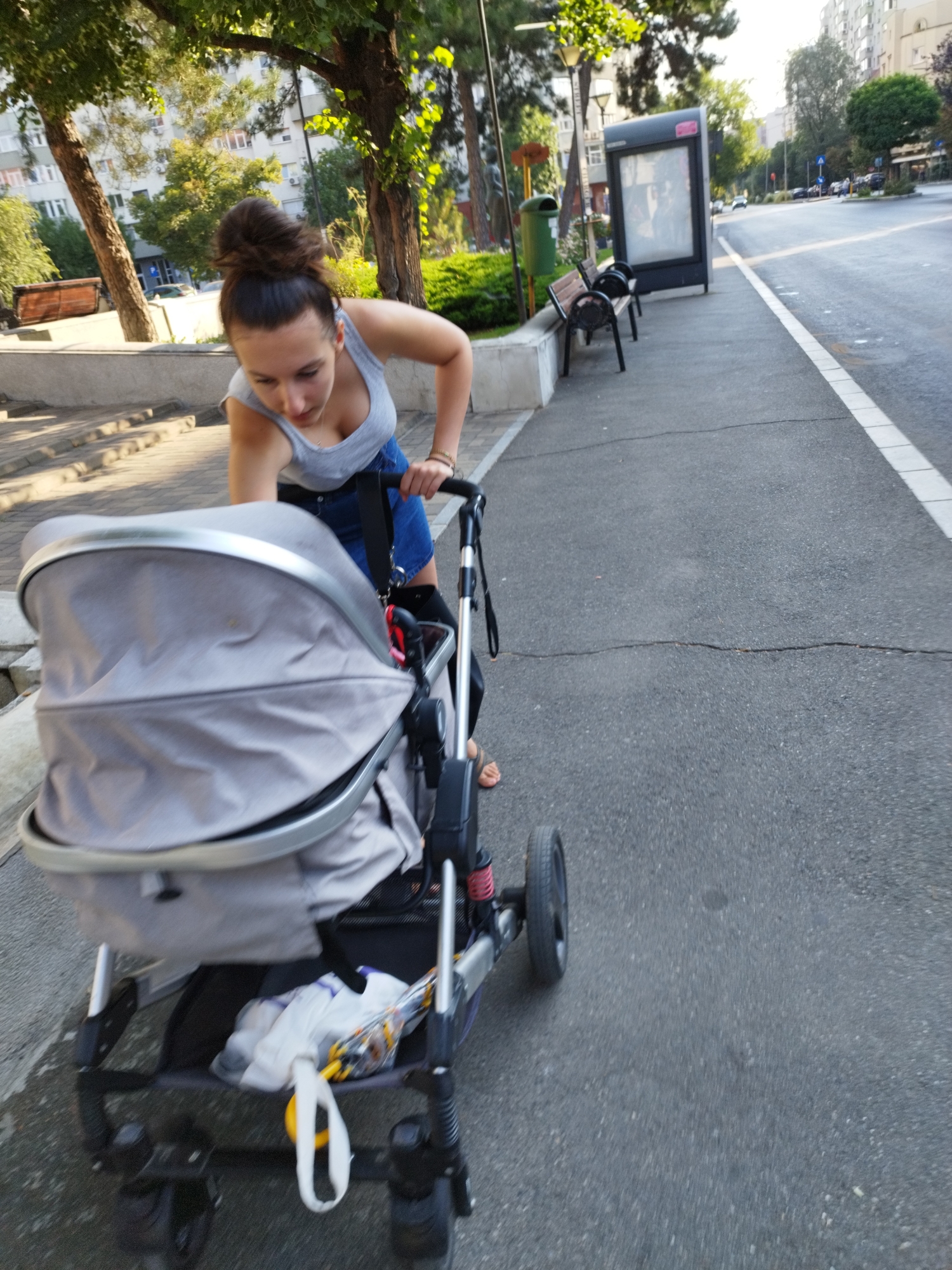 In Bucharest, Romania, a young woman with her hair in a bun, wearing a gray tank top and a denim skirt, pushes a gray stroller. The stroller has a large canopy, and a white bag with yellow and orange items is visible in the lower basket. The woman is leaning forward, with her right hand on the stroller handle, and her left hand appears to be holding it as well. She is wearing sandals. The scene takes place on a sidewalk next to a street, with trees and buildings in the background. There are also park benches and a bus stop sign visible. The lighting suggests it is daytime, possibly late afternoon.