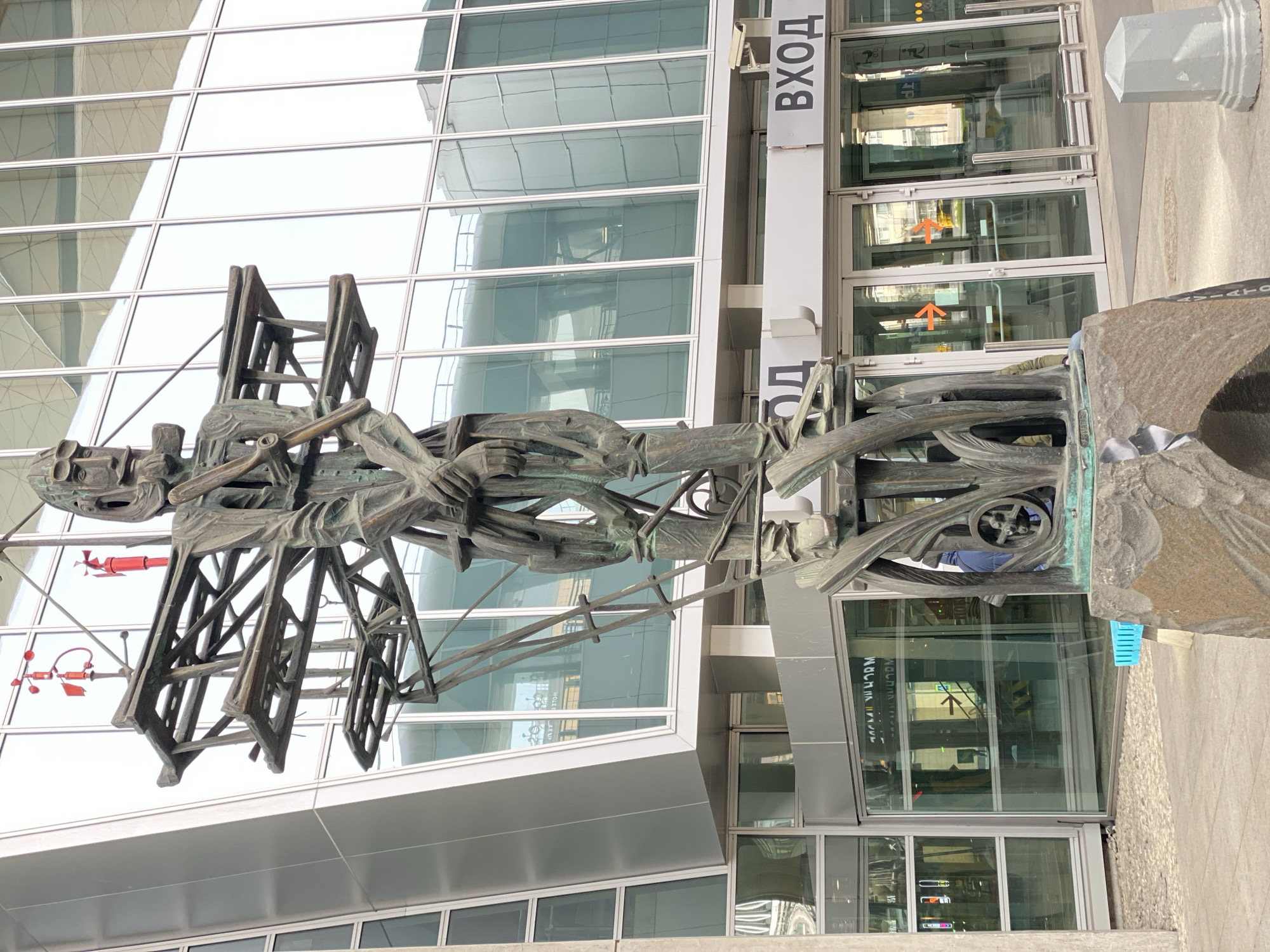 The image shows a bronze sculpture in front of a modern building with glass windows and an entrance labeled "ВХОД," which means "Entrance" in Russian. The sculpture depicts a person holding what looks like a ladder. The sculpture is located in Saint Petersburg, Russia, outside the Pulkovo Airport.