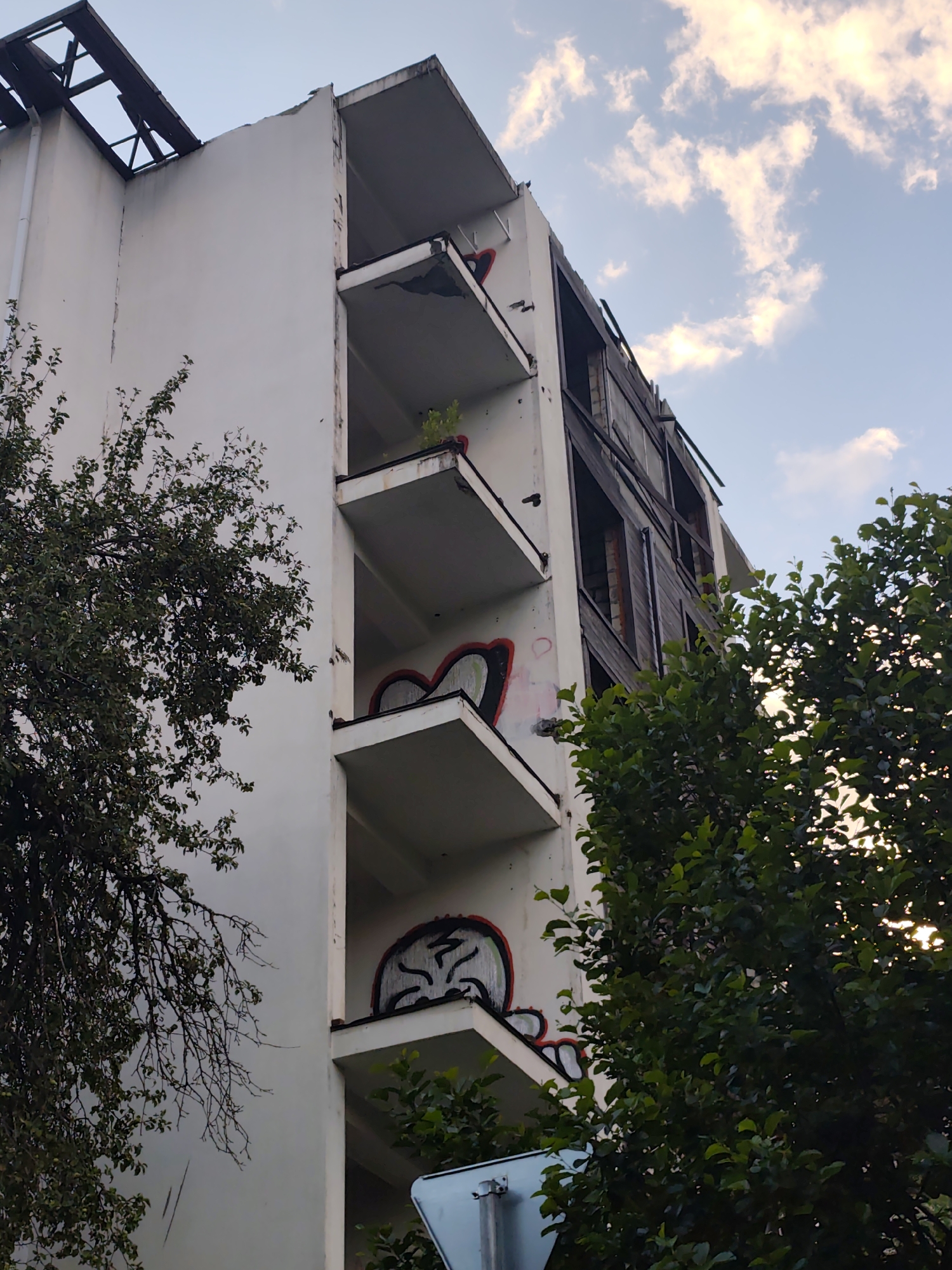 This media file captures a weathered, abandoned multi-story building, likely an apartment complex, in Krasnaya Polyana, Russia. The building exhibits signs of decay, with damaged balconies, exposed structural elements at the top, and wooden boards covering some of the windows. Graffiti art, primarily in red and black, adorns several balconies, depicting stylized faces or abstract shapes. Green foliage from surrounding trees partially obscures the structure, suggesting a natural or overgrown environment. The sky above is partly cloudy, with visible patches of blue and white clouds, hinting at daytime, possibly late afternoon or early evening due to the soft lighting. The overall mood conveyed is one of neglect and urban decay, juxtaposed with the resilience of nature. No people are visible, and no text is discernible in the image.