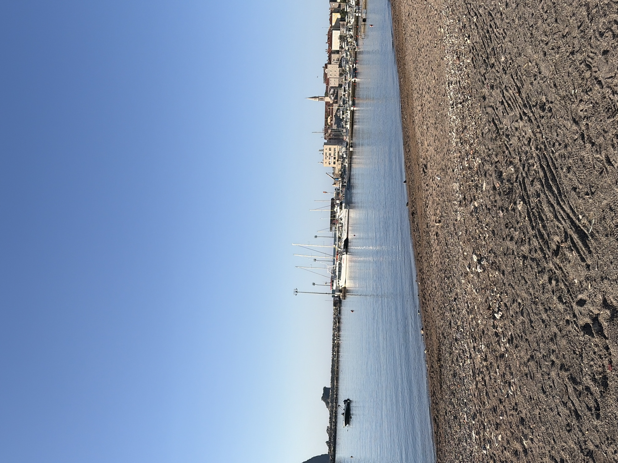 The image depicts a coastal scene in Budva, Montenegro. On the left is a clear, bright blue sky. Adjacent to the sky is a body of water with boats docked. Along the right of the water is a shore with textured sand. In the distance, buildings can be seen.