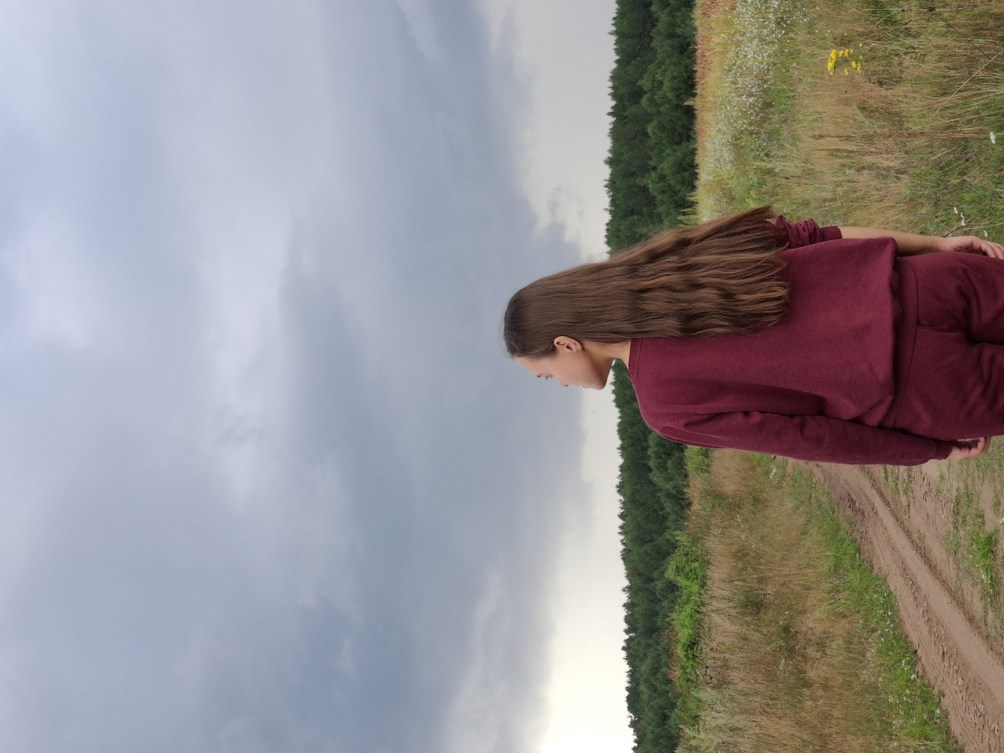 In Plishkino, Russia, a young person with long, brown hair, viewed from behind, stands on a dirt path in a field. They are wearing a burgundy tracksuit. The path leads towards a dense line of dark green trees in the background. The sky above is overcast with heavy, gray clouds, suggesting impending rain or a storm. The field is filled with dry, yellowing grass and some small white and yellow wildflowers.