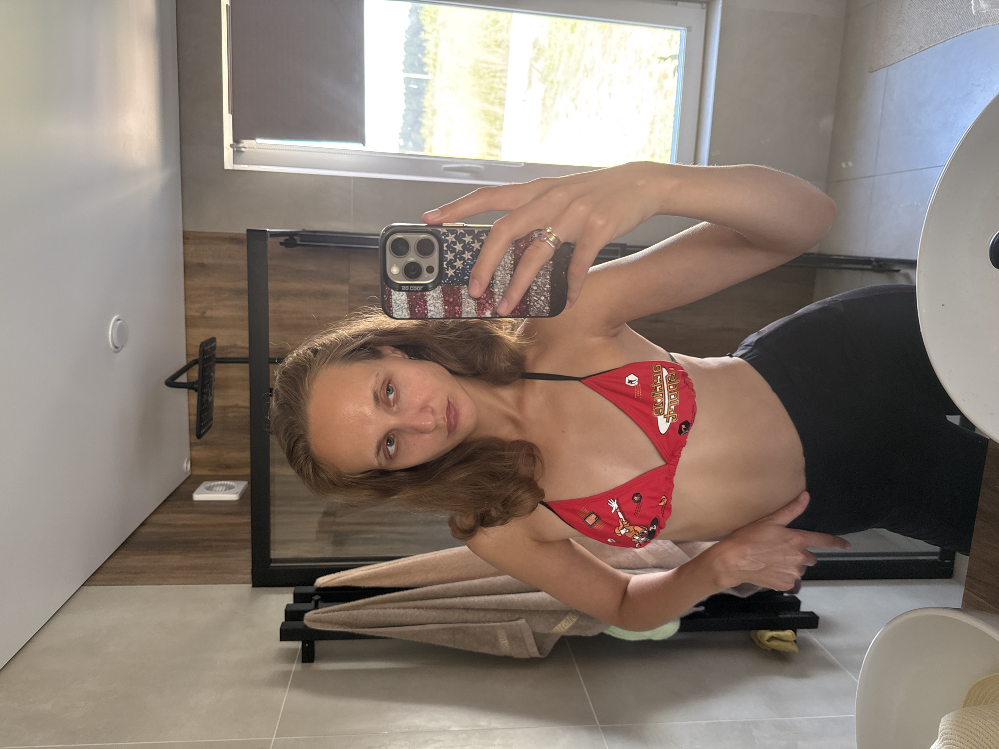 A young woman is taking a mirror selfie in a modern bathroom. She is wearing a red bikini top with a cartoon pattern and black leggings. The bathroom features beige tiled walls, a wooden accent wall behind the shower, and a large window with a view of a grassy field and trees. There are towels hanging on a rack, a shower head, and a sink visible. She is holding a smartphone with an American flag patterned case, and wearing a ring on her finger. The woman's expression is neutral.