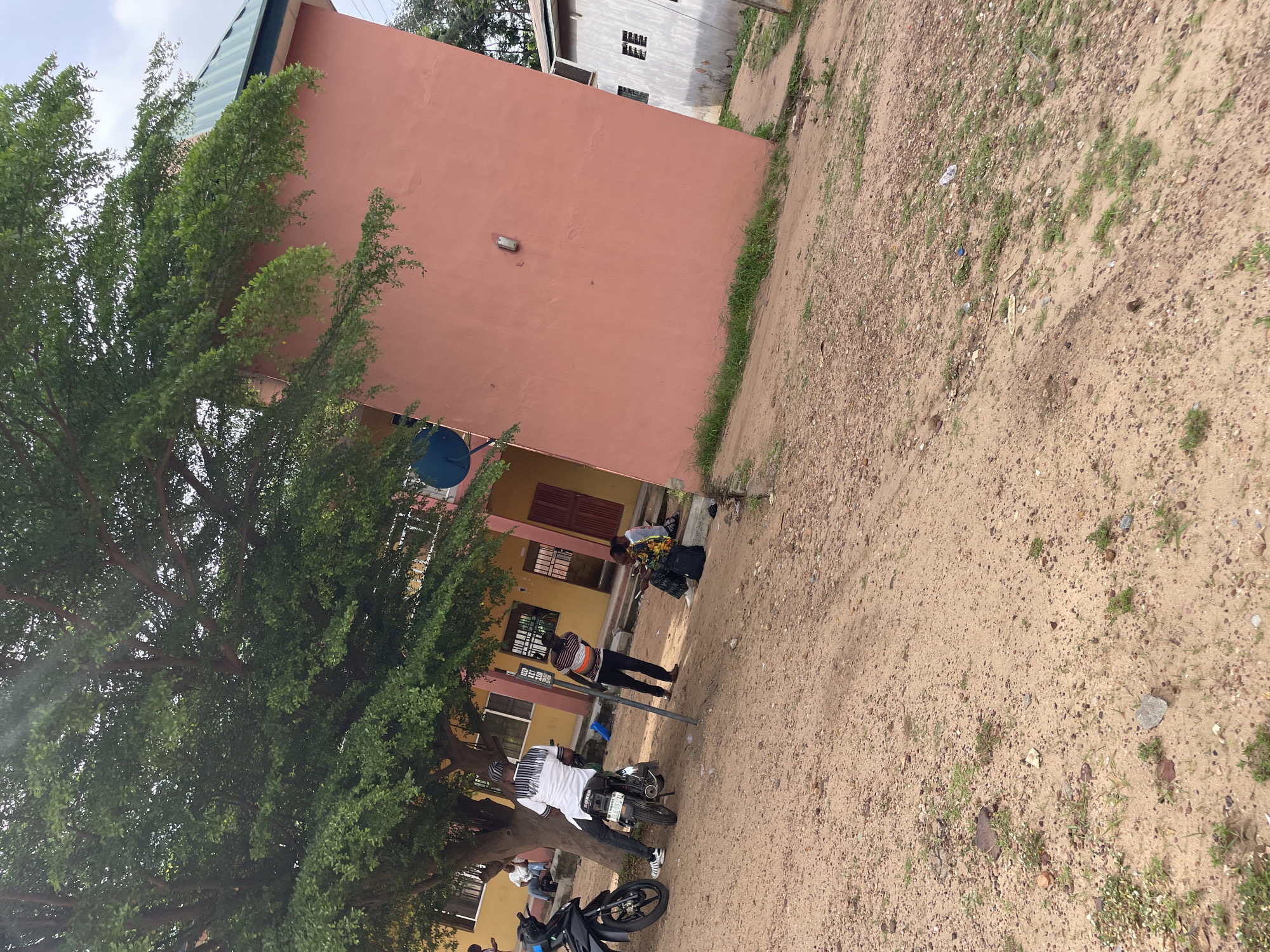 The image depicts an outdoor scene in Lokoja, Nigeria. In the foreground, a sandy, uneven ground is visible with scattered small stones and patches of sparse green grass.
Dominating the left side of the image is a large tree with lush green foliage. To the left of the tree, a black motorcycle is parked. Behind the motorcycle, a person is seated on another motorcycle, facing away from the camera, wearing a white shirt with black decorative patterns on the shoulders and dark trousers. They are near the base of the tree.
Behind the tree and the motorcycles is a building with a yellow facade. Several windows with white frames are visible on this building. To the right of the yellow building, a larger, salmon-pink colored building extends into the frame. This building has a green, slanted roof. A dark blue satellite dish is attached to the upper part of the salmon-pink building, near the corner where it meets the yellow building.
Further to the right, a lighter colored building is partially visible, with a white facade and windows featuring barred openings.
In front of the buildings, a few individuals are present. Two people are seated on the ground near the salmon-pink building, one holding a dark bag. Another person, wearing a striped t-shirt and dark trousers, stands between the motorcycles and the seated individuals, looking towards the building. A person is also seen walking near the yellow building.
The overall impression is a somewhat disheveled outdoor area adjacent to buildings, possibly a school or residential compound, with evidence of daily activities like motorcycle use and people gathering. The lighting suggests it is daytime, with a cloudy sky.
