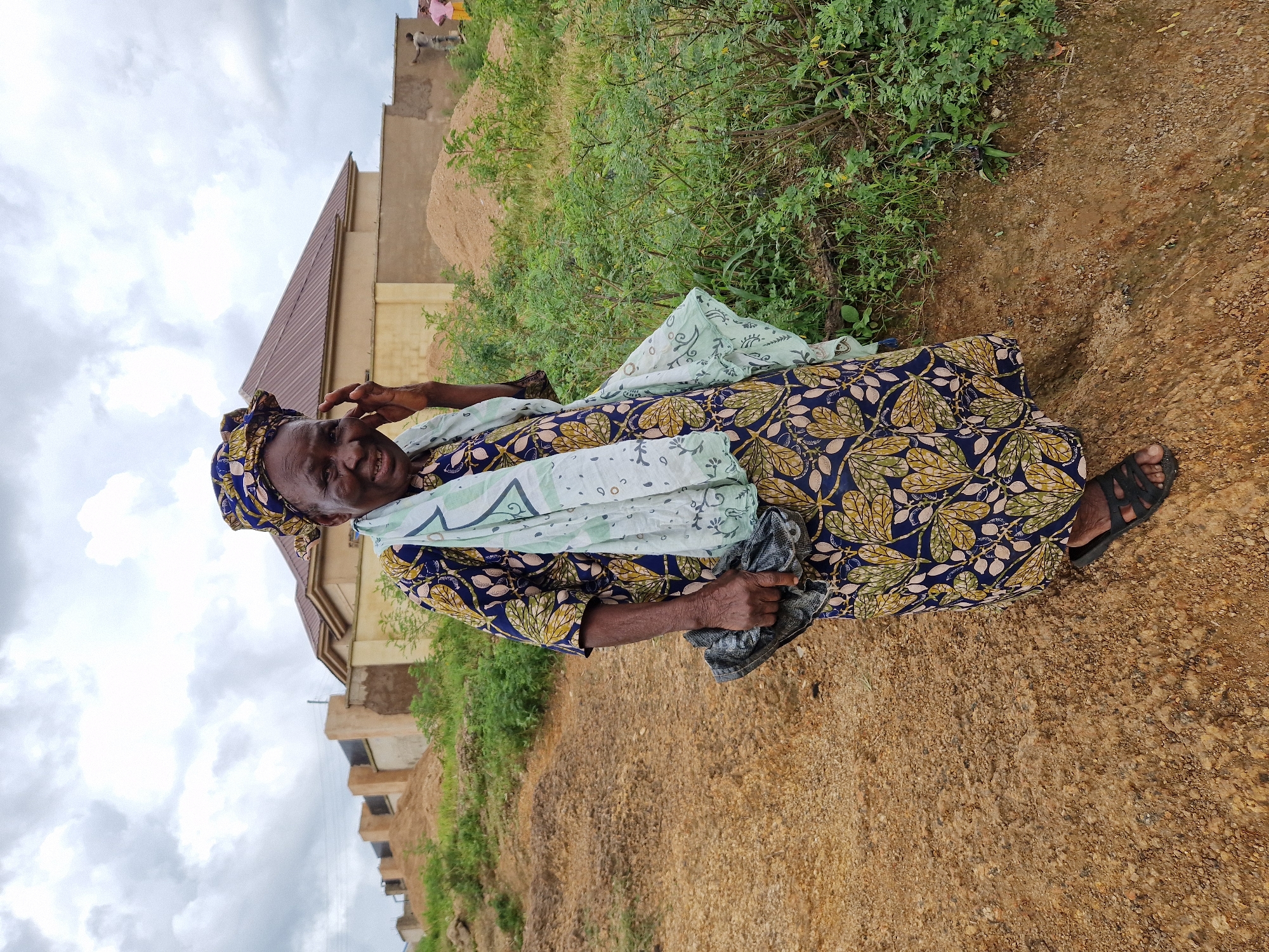 A full-shot, outdoor, eye-level, medium-light shot depicts an elderly Nigerian woman standing in a dirt lot, with a building in the background. The woman is wearing a blue, gold, and green patterned dress with a matching headscarf and a light green scarf draped over her shoulders. She is smiling, with her right hand touching her face, and holding a black bag in her left hand. She is wearing black sandals. The background shows a large, light brown building with a brown roof, behind a beige wall. There are piles of sand and green vegetation surrounding the building and the woman. The sky is overcast and cloudy. The image appears to have been taken in a rural or developing area of Jos, Nigeria, given the construction, unpaved ground, and vegetation.