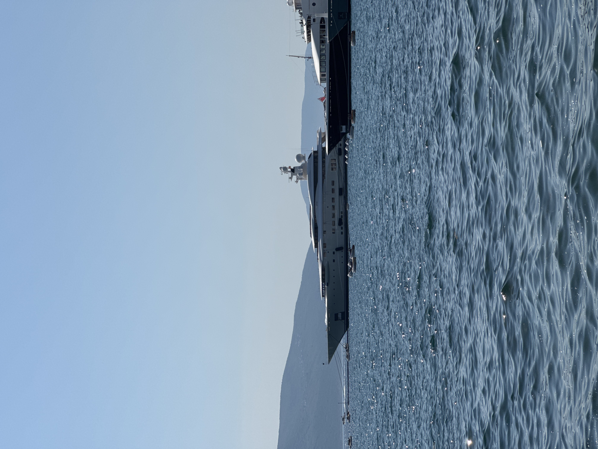 A serene view of the Tivat bay in Montenegro on a clear day. The foreground is dominated by the deep blue, rippling water of the bay, with sunlight glinting off the surface, creating a sparkling effect. In the mid-ground, two large yachts are anchored. The larger yacht on the left is predominantly white, with multiple decks and a sleek, modern design, featuring prominent radar domes and other equipment on its upper levels. To its right, a second, darker-hulled yacht is visible, also appearing to be a luxury vessel. Smaller boats, possibly tenders or dinghies, are scattered around the yachts. In the background, lush green mountains form a scenic backdrop to the bay, meeting the clear blue sky. The overall scene depicts a tranquil and picturesque maritime setting.