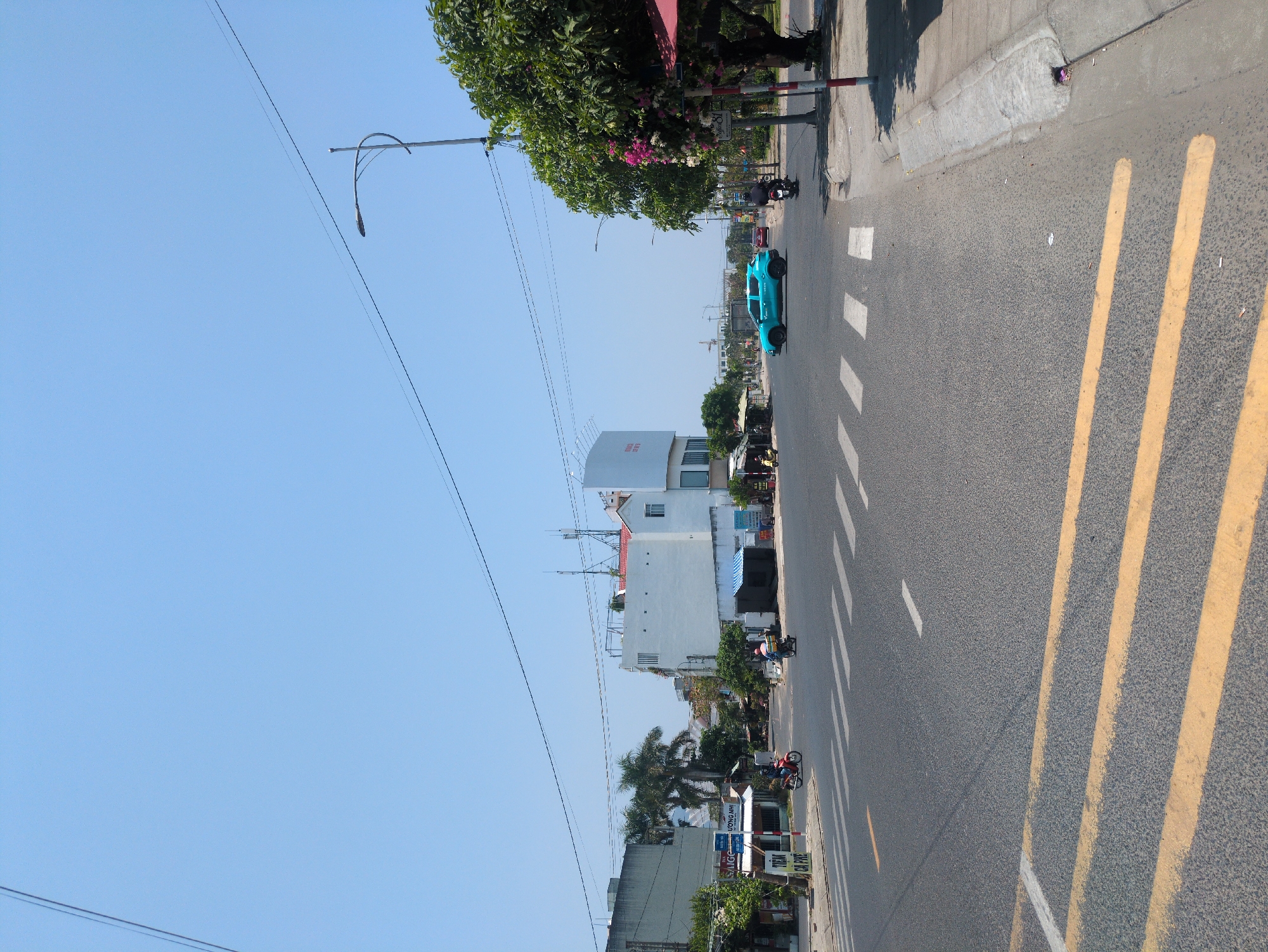 The image depicts a street scene in Tuy Hòa, Vietnam. A light blue pickup truck is driving on the right side of the road, heading away from the camera. There are two motorcycles in the distance. The street has yellow double lines indicating a no-passing zone and white stripes marking a pedestrian crossing. Buildings line the left side of the street, with a modern white building closer to the camera and older, more traditional structures farther away. A street light is visible on the left. Power lines stretch across the sky. The sky is clear and blue. Trees and vegetation are present on both sides of the street.