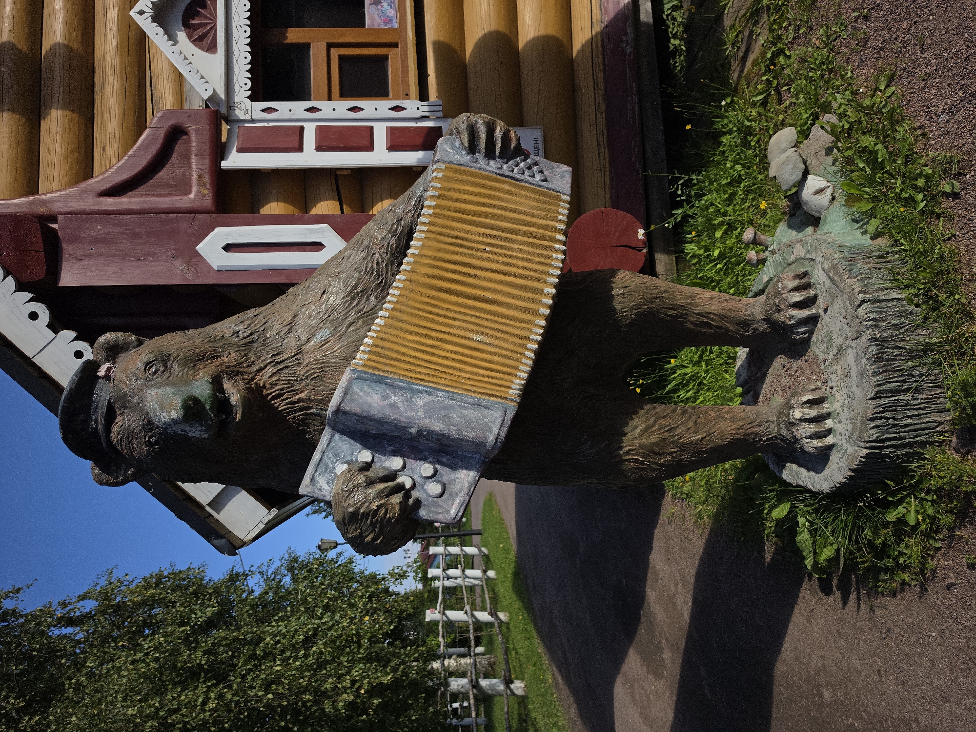 A statue of a brown bear wearing a black cap stands on a tree stump, holding an accordion. The bear statue is located in front of a wooden building with red and white decorative trim. The building appears to be a shop or restaurant, typical of Russian folk architecture. The environment is outdoors, with a clear blue sky, green trees, grass, and a paved path. A white wooden fence is visible in the background. This scene is located in the city of Peterhof, Russia, likely within its park complex or a village area. The lighting suggests it is a sunny day.