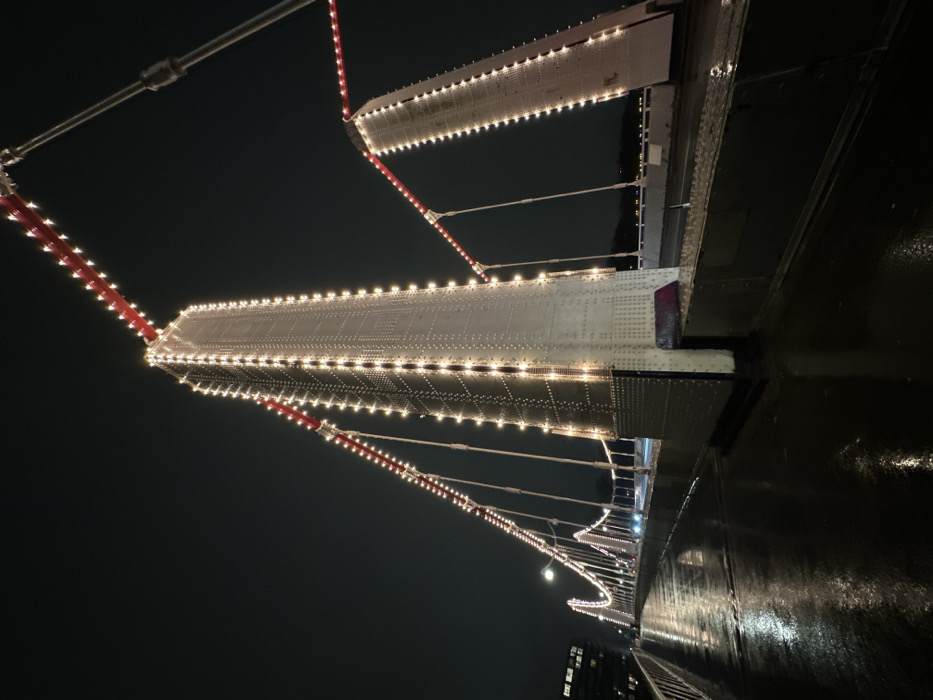 An upward-angled shot captures the structure of a bridge illuminated at night. Rows of lights trace the edges of the bridge's supports and suspension cables. The bridge's reflective surface gives a slight mirroring effect to the lights. Dark sky serves as the background, providing contrast to the illuminated bridge. A building with illuminated windows appears at the end of the bridge.