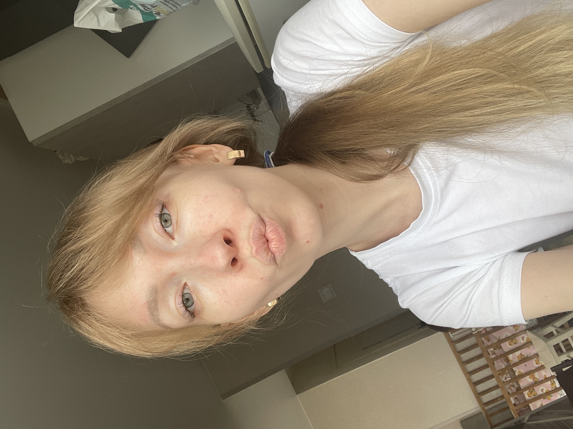 A pale-skinned woman with long, blonde hair and light-colored eyes looks at the camera with pursed lips. She is wearing a white t-shirt. The background shows a room with gray walls and a wooden crib. The location information provided indicates this might be in Perm, Russia.