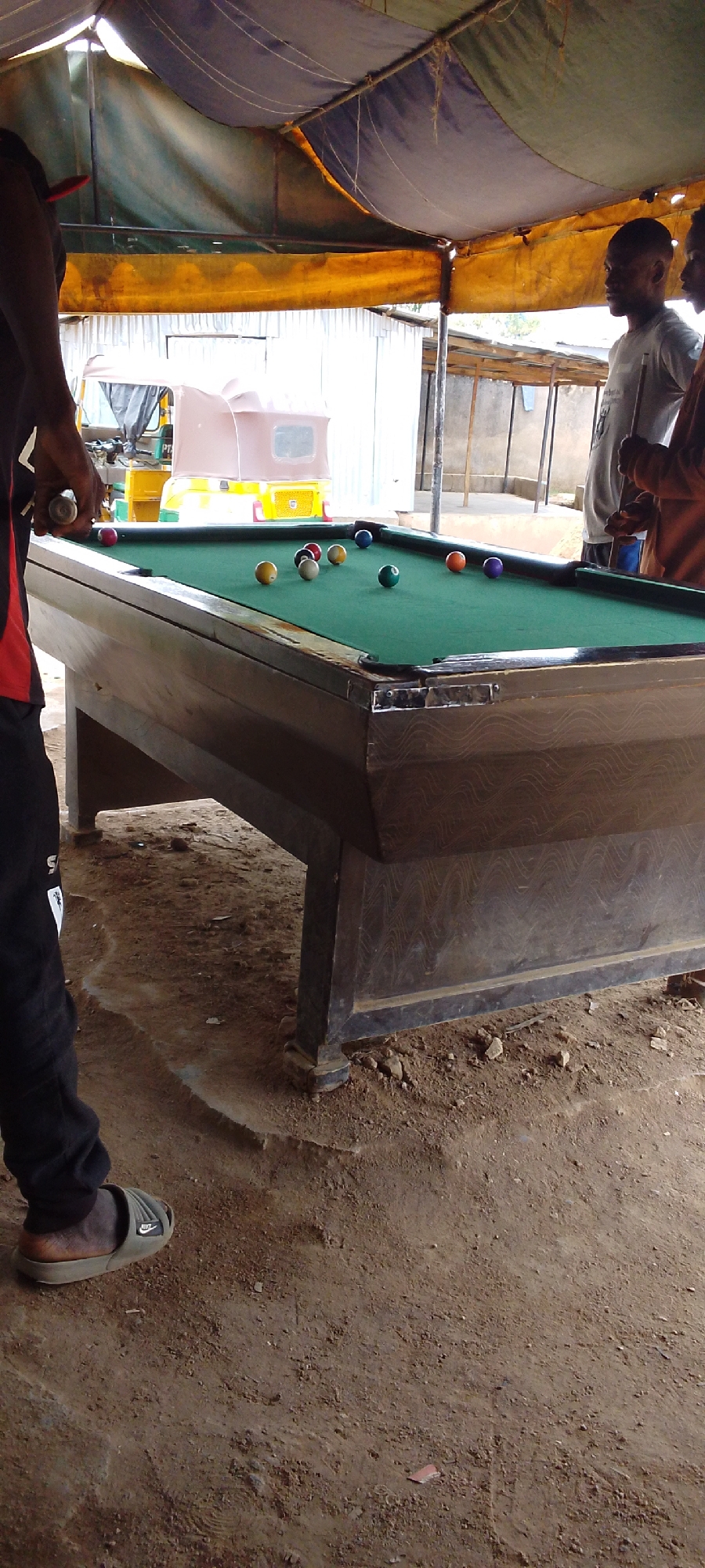 The image captures an outdoor pool hall scene in Jos, Nigeria. The foreground features a worn billiards table with multiple balls scattered across its green surface. A man is visible on the left side of the frame, preparing to take a shot. He is wearing a red shirt with black pants and Nike slides. Two other men stand to the right of the pool table, observing. The hall is covered with a yellow, green, and blue tarp. Behind the pool table, there is a three-wheeled vehicle, or a "Keke Napep", parked outdoors. The ground is bare earth and shows signs of wear and tear. Buildings and open structures are visible in the background, indicating an urban setting.