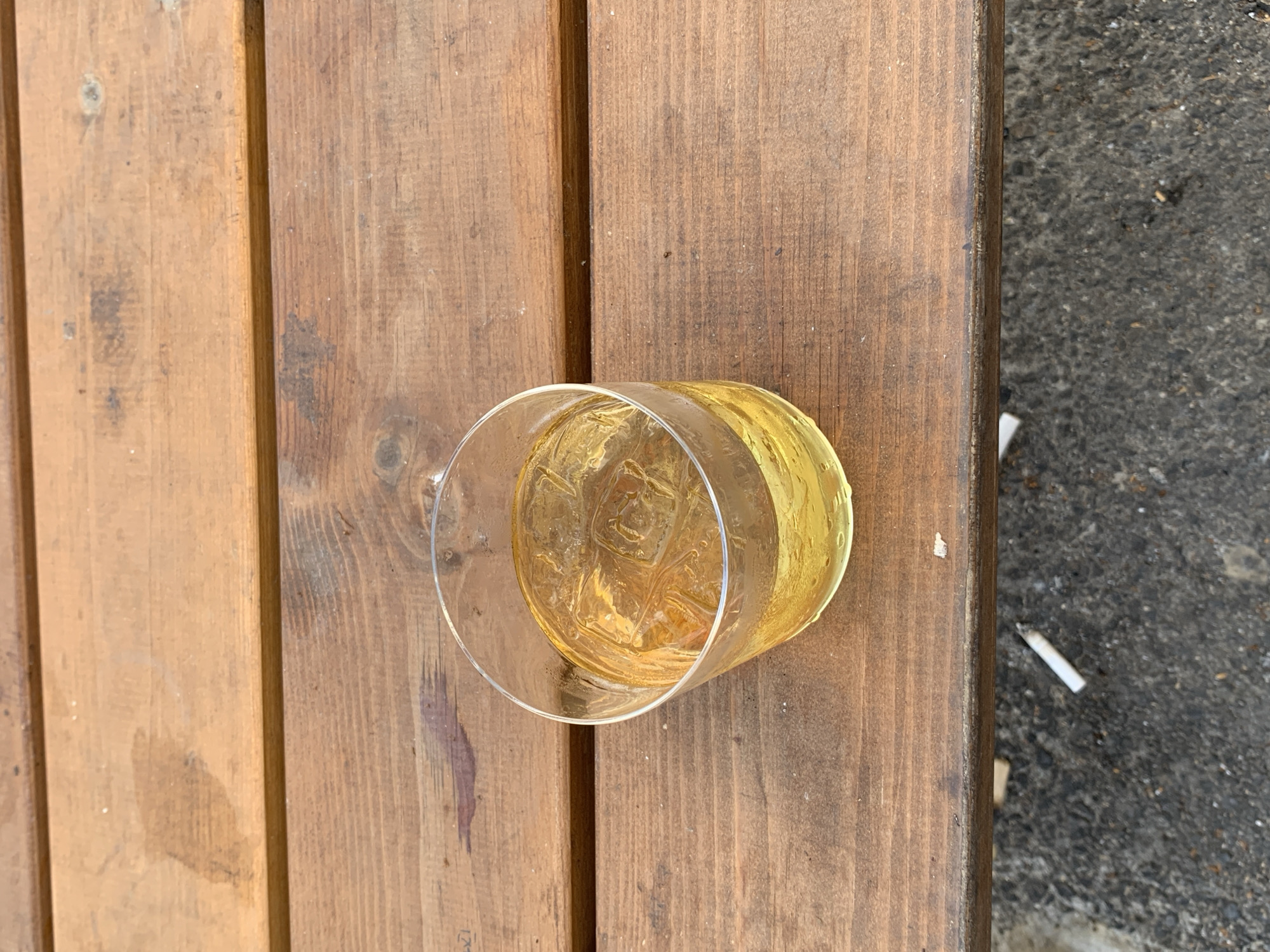A clear glass of amber liquid with ice cubes rests on a wooden surface made of several planks. The wooden surface appears to be part of outdoor furniture, possibly a bench or table. Adjacent to the wood is a gray, textured surface, possibly pavement, on which discarded cigarette butts are visible. The setting appears to be outdoors. While identifying the exact location as Tel Aviv, Israel, is not possible with only this image, the presence of cigarette butts and outdoor furniture is consistent with public spaces found in many urban areas. No people are visible.