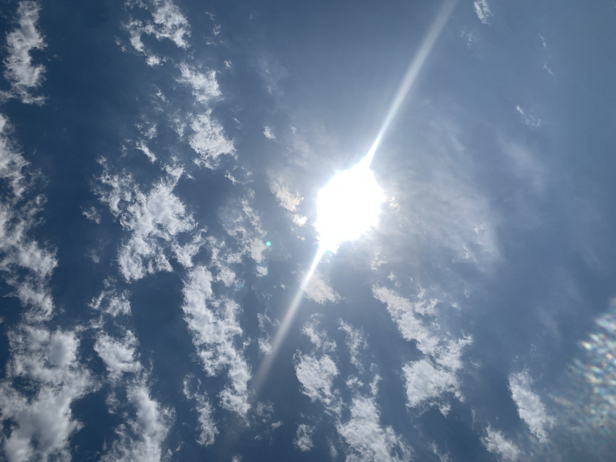 This is a photograph of a bright sun in a blue sky with scattered white clouds. The sun is positioned in the center of the image, with rays of light emanating from it. Some of the clouds are thin and wispy, while others are thicker and more defined. There are some lens flares visible in the bottom left corner of the image, appearing as colorful circles. The overall impression is one of a clear, sunny day. The scene is not specific to any particular location in the city of Khazar Raion, Azerbaijan, as it depicts only the sky and sun.