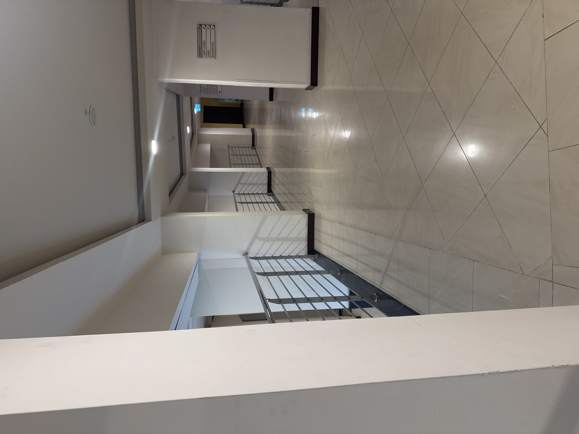 This is a photograph of a brightly lit, modern indoor hallway. The hallway features a polished tile floor with a diamond pattern. On the left side of the image, a white column is prominent, and beyond it, a metal railing with horizontal bars overlooks a lower level. The ceiling is a complex structure with recessed lighting and decorative elements. On the right side, a white pillar supports a sign with directions to "FAR EAST", "LOBBY LOUNGE", "LOBBY", and "IN DOOR POOL". A dark doorway with an illuminated green exit sign is visible in the distance at the end of the hallway. The overall impression is one of cleanliness and contemporary design. The location is not specified to be in Aksu, Turkey.