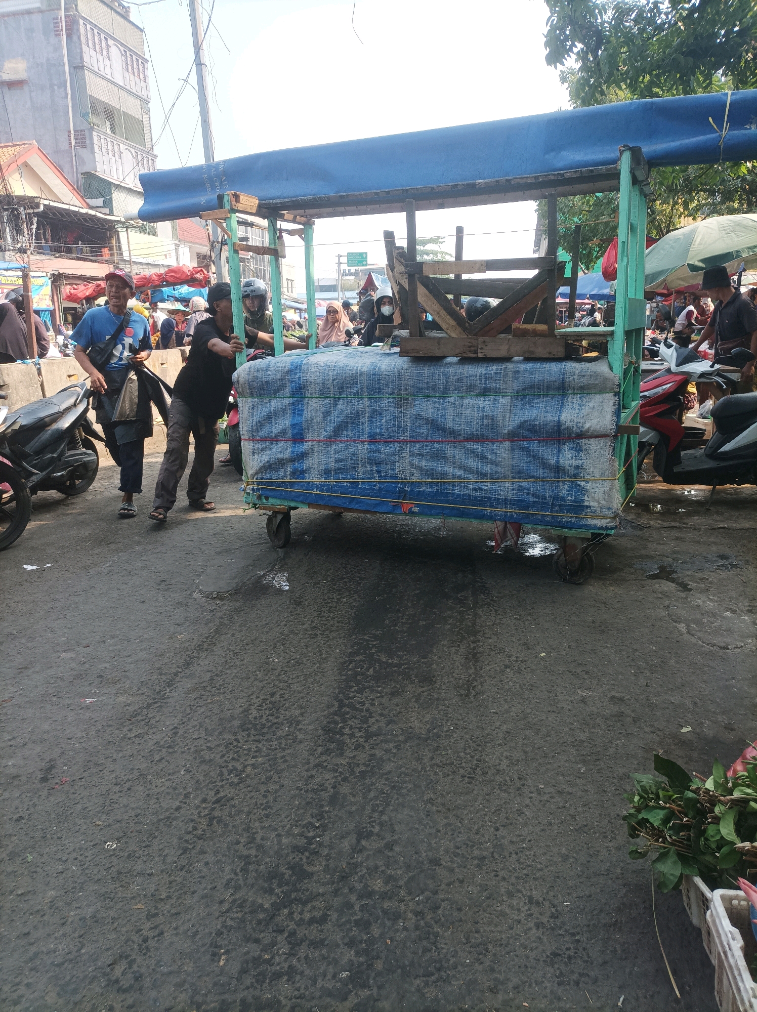 In a street scene in South Jakarta, Indonesia, a street vendor is seen pushing a cart laden with goods wrapped in blue tarp. The cart, constructed with a wooden frame and supported by wheels, is positioned on an asphalt road. A man in a blue t-shirt and dark pants stands to the left, holding a plastic bag. Another person in dark clothing is on the right, also pushing the cart. Behind them, various market stalls and buildings are visible, with trees and electrical wires overhead. Several motorcycles are parked in the vicinity. The overall environment suggests a bustling market atmosphere.