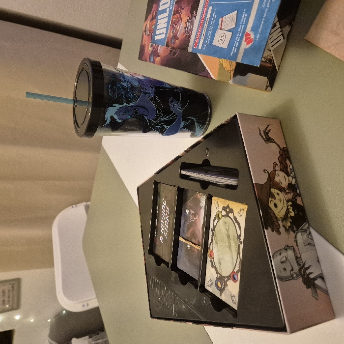 A black cup with a blue straw sits next to a game box titled "Unlo." A board game box with the title "A Inside Story" is open, revealing card slots and cards. The game box has illustrations on its side, including a character with a wide-brimmed hat and a smaller figure resembling a robot. The items rest on a white surface, which appears to be a table.