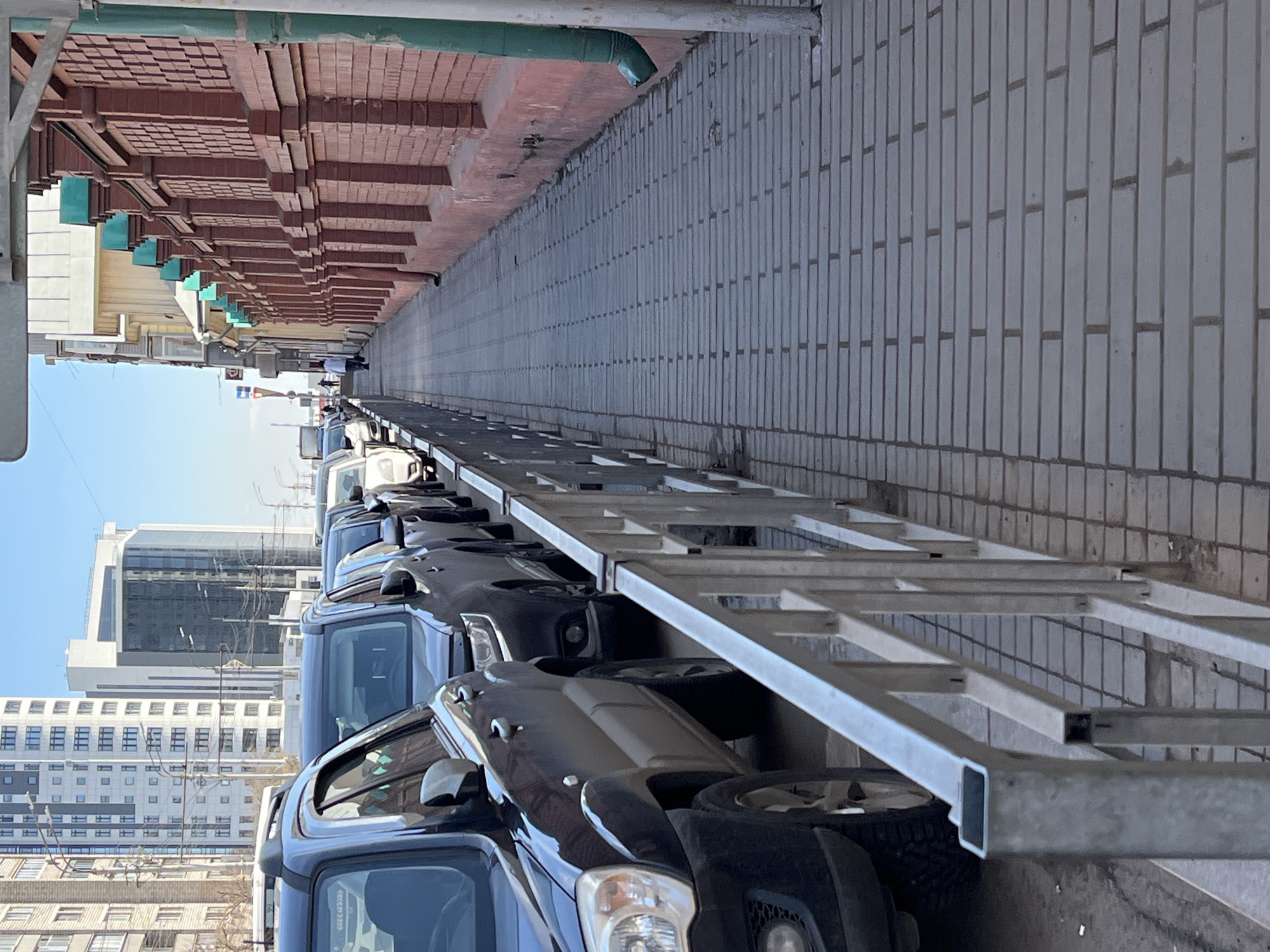 The image, when viewed upright, presents an urban street or alleyway under bright daylight. A long, narrow paved area, likely a sidewalk or service road, stretches into the distance, creating a strong sense of perspective.
On the left side of this paved area, a line of dark-colored vehicles, primarily SUVs or trucks, is parked bumper-to-bumper. Some of these vehicles appear to be positioned on a multi-level metal structure, possibly a car carrier or a display rack, given the silver-colored metal beams and platforms visible above and between them. The front headlights and tires of several vehicles are clearly visible.
To the right of the parked vehicles, a tall, light grey brick wall with a distinctive vertical bonding pattern extends along the length of the pathway. Above this wall, a long, older building with reddish-brown brickwork forms the boundary on the far right. This building features decorative architectural elements, including recessed sections and what appear to be small, turquoise-colored awnings or projections lining its facade. A green drainpipe is visible along the upper edge of this building.
In the far background, under a clear, bright blue sky, several modern high-rise buildings with glass facades and white concrete elements can be seen, contrasting with the older brick architecture in the midground. A few small, indistinct figures are visible in the distance along the paved path. The scene is brightly lit, indicating a sunny day. No discernible text is visible in the image.