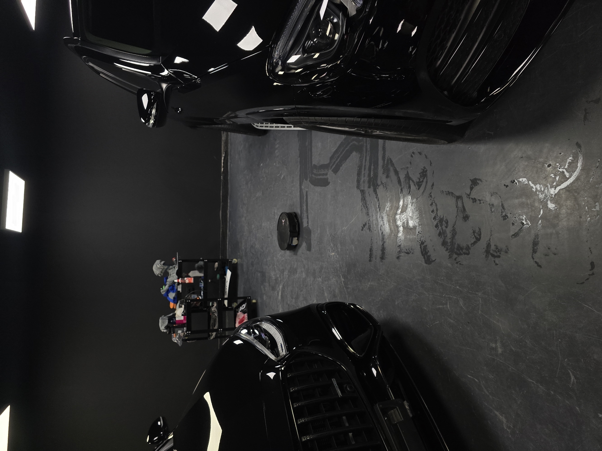 The image depicts a dimly lit indoor space, possibly a garage or workshop, with a dark grey or black wall as the backdrop. Two black luxury cars are parked side-by-side, with the front of one vehicle on the left and the side of another on the right, suggesting they are positioned in a way that their fronts are not facing the viewer directly. A black robotic vacuum cleaner is visible on the floor between the cars. To the left, a rolling cart is laden with various cleaning supplies, including bottles and cloths, indicating a car detailing or maintenance area. The floor appears to be concrete, with some wet patches and streaks, possibly from cleaning activities. The lighting comes from overhead square fixtures, casting reflections on the polished surfaces of the cars and the floor. There are no people visible in the scene. The image does not provide any specific information or visual cues that would allow for an identification of the location as Ruse, Bulgaria.
