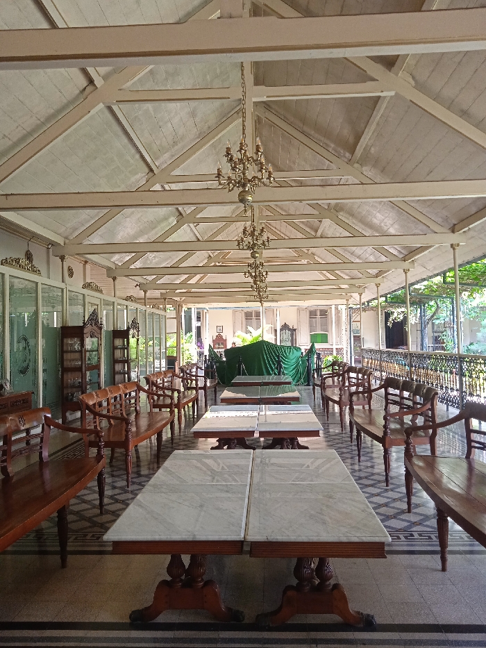 The image shows a long, covered veranda in a building located at -7.566 latitude and 110.823 longitude in Surakarta, Indonesia. The veranda has a white, wood-beamed ceiling with two chandeliers hanging from the beams. The floor is tiled in a black and white geometric pattern. There are four long, wooden benches with padded seats on each side of the veranda. In the middle of the veranda, two long, white marble tables with a brown, wooden base are placed side-by-side. The tables are empty. Through the windows and doors at the end of the veranda, one can see lush, green trees. There are a number of ornate cabinets and chairs in the background, all made of wood. The entire structure is built in a traditional Indonesian style. There are no people present.