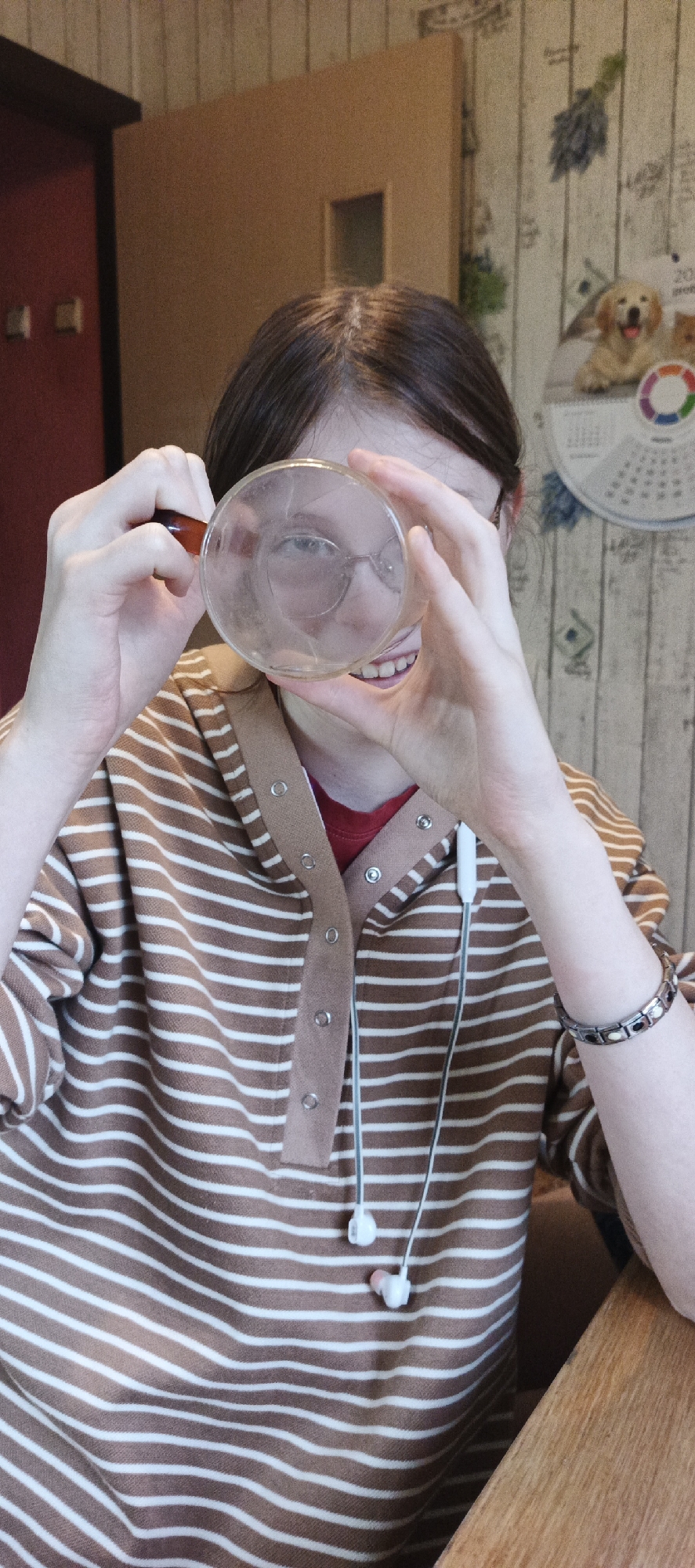 An eye-level shot captures a person indoors, holding a clear glass object up to their face with both hands, looking through it. The person has dark brown hair and is wearing glasses, which are visible and slightly distorted through the glass. A wide smile is evident on their face. They are dressed in a brown and white striped long-sleeved top with a button-up placket, worn over a red t-shirt. White wired earbuds with pink tips hang around their neck. A metal bracelet adorns their left wrist.
The background shows a domestic setting. To the left, a dark reddish-brown doorframe is visible against a light brown wall or door. To the right, a light-colored wall with faint patterns features a calendar hanging on it. The calendar displays a picture of a golden retriever, a circular color wheel, and partially visible text, including "202" (likely part of a year) and "gree". The person is seated at a wooden table, part of which is visible in the lower right. The overall impression is one of a playful or curious moment.