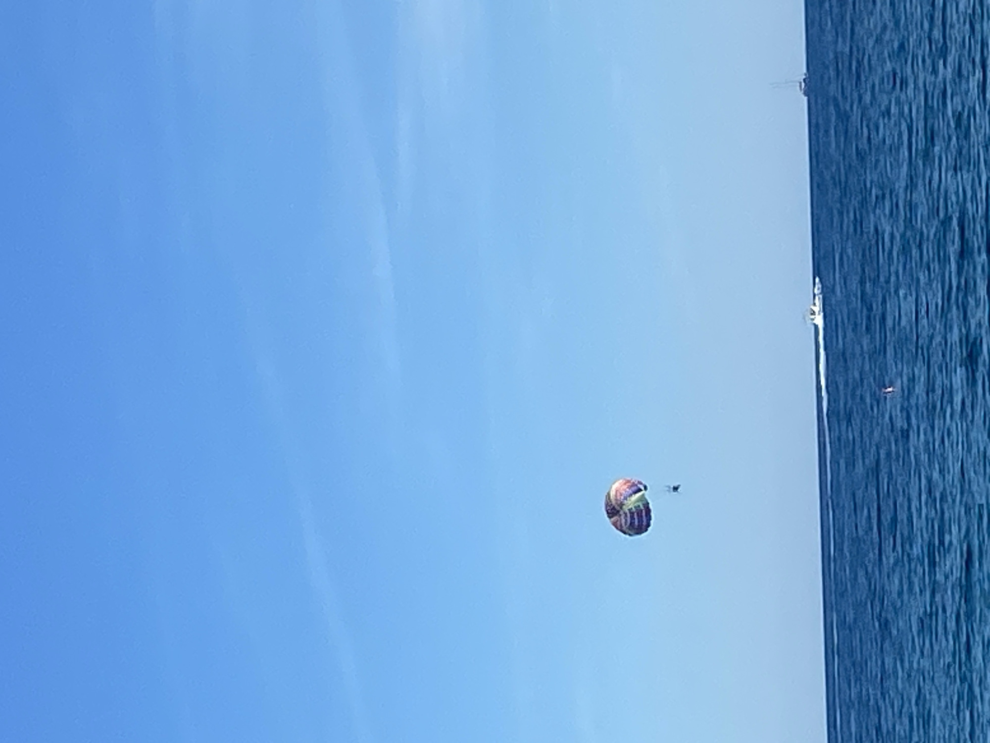 A medium-range shot captures a parasailer suspended in the air above the coastal waters of Boreti, Montenegro. The parasail is multicolored. A small boat is visible in the water. The sky is clear and blue.