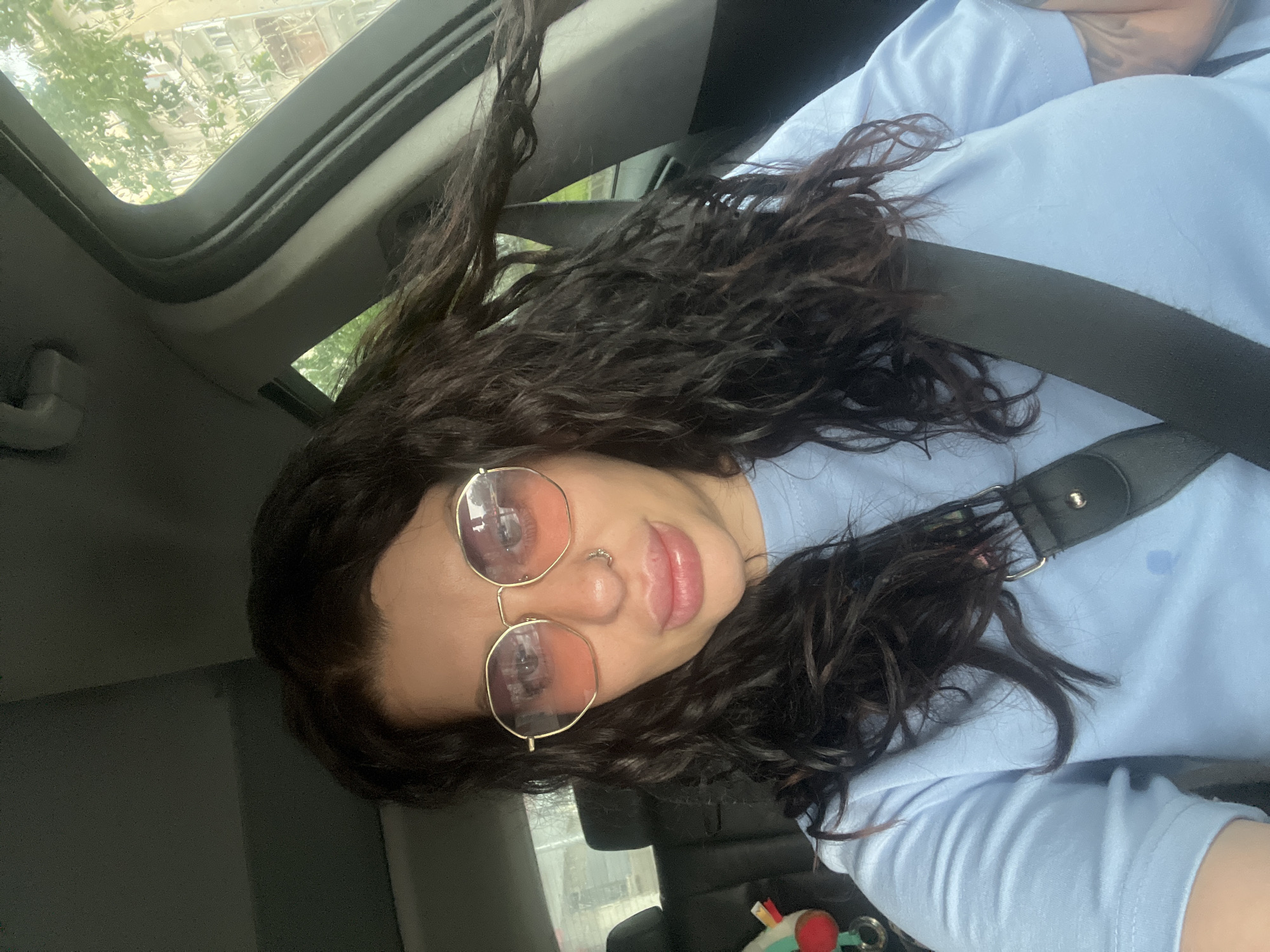 The media file presents a rotated selfie of a person inside a car, looking directly at the camera. The subject has long, dark, curly hair and is wearing a light blue t-shirt. A dark seatbelt is diagonally across their chest. They are wearing round, gold-framed sunglasses with reflective, pink-tinted lenses, and a septum nose piercing is visible. Their lips are slightly parted, suggesting a neutral or soft expression.
Through the car's open sunroof, blurred green foliage from trees and indistinct buildings are visible against a bright sky, indicating that the photo was taken during daylight hours. The interior of the car appears dark in contrast to the bright exterior light, with parts of the headliner and pillars visible. Faint tattoo outlines can be seen on the person's arm or shoulder beneath the t-shirt. In the lower right corner of the image, some small objects, including a red cap and a white and green item, are partially visible in what appears to be a car console or cupholder. The scene captures a casual moment within a vehicle, possibly while driving or parked. No visible text is present in the image.