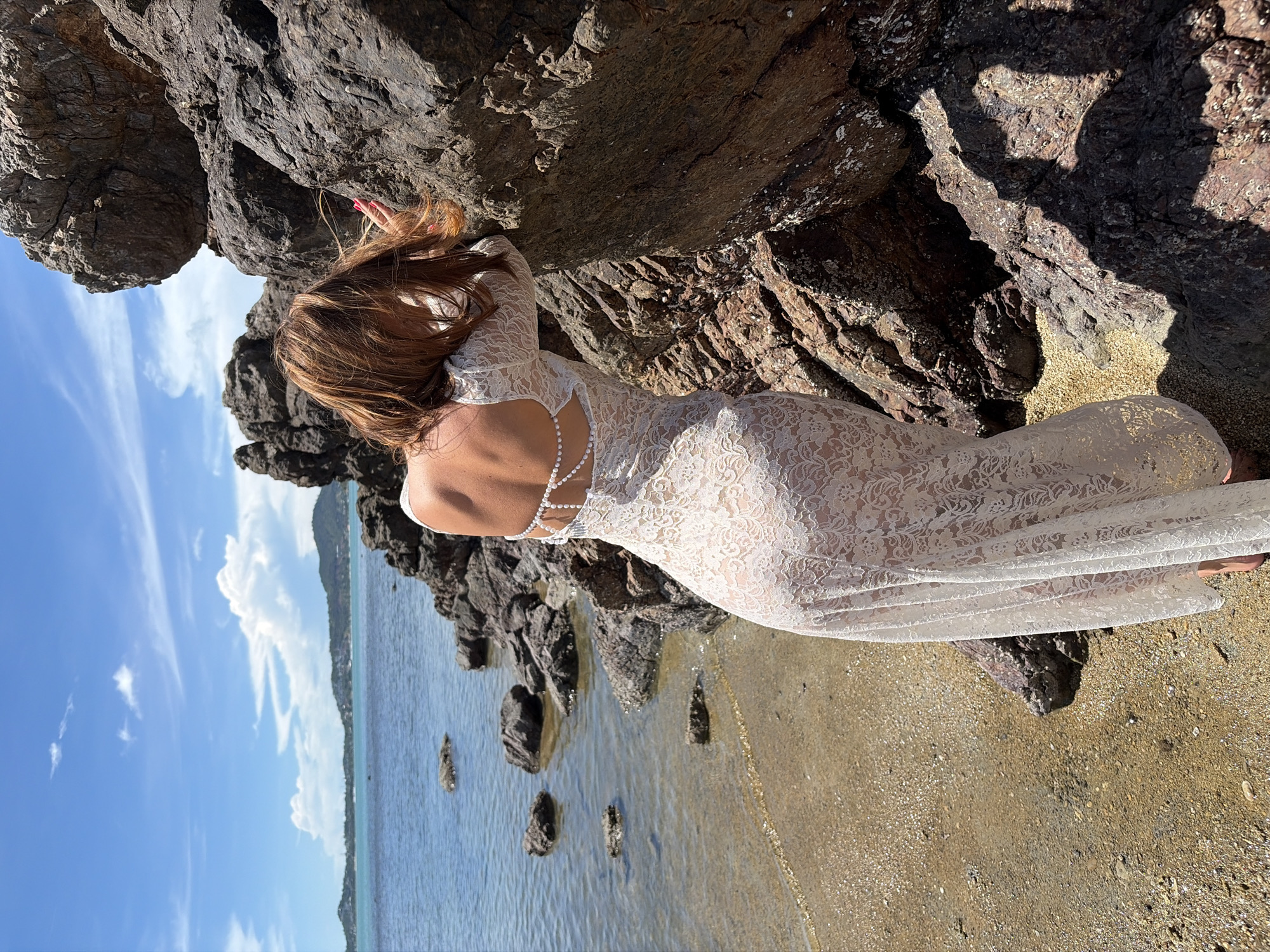 A woman with long, reddish-brown hair, viewed from the back, leans against dark, jagged rocks near the ocean. She is wearing a white lace dress with an open back, adorned with a delicate pearl-like embellishment. The dress is long and form-fitting, with intricate floral lace patterns visible through the fabric. Her right hand is resting on the rocks, and her left arm is bent. The scene appears to be set on a rocky shore with some sand. The ocean is calm and blue, reflecting the bright sky, which is dotted with white, wispy clouds. In the distance, a faint outline of mountains or hills can be seen. The lighting suggests it is daytime, likely sunny with some shadows cast by the rocks. The overall mood is serene and peaceful, with a focus on the natural beauty of the location and the woman's pose. There is no visible text in the image. The location context is Baan Bo Phut, Thailand.