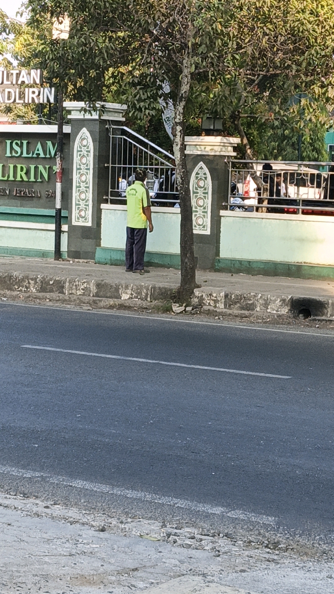 In Kuwasen, Indonesia, a person stands on a sidewalk in front of a building with a sign that reads "ISLAM" and "ADLIRIN" with "EN JEPARA 694" below it. The building is painted green and white with decorative elements. The person is wearing a bright green shirt and dark pants. A tree is positioned between the person and the road. The road in the foreground appears to be asphalt.