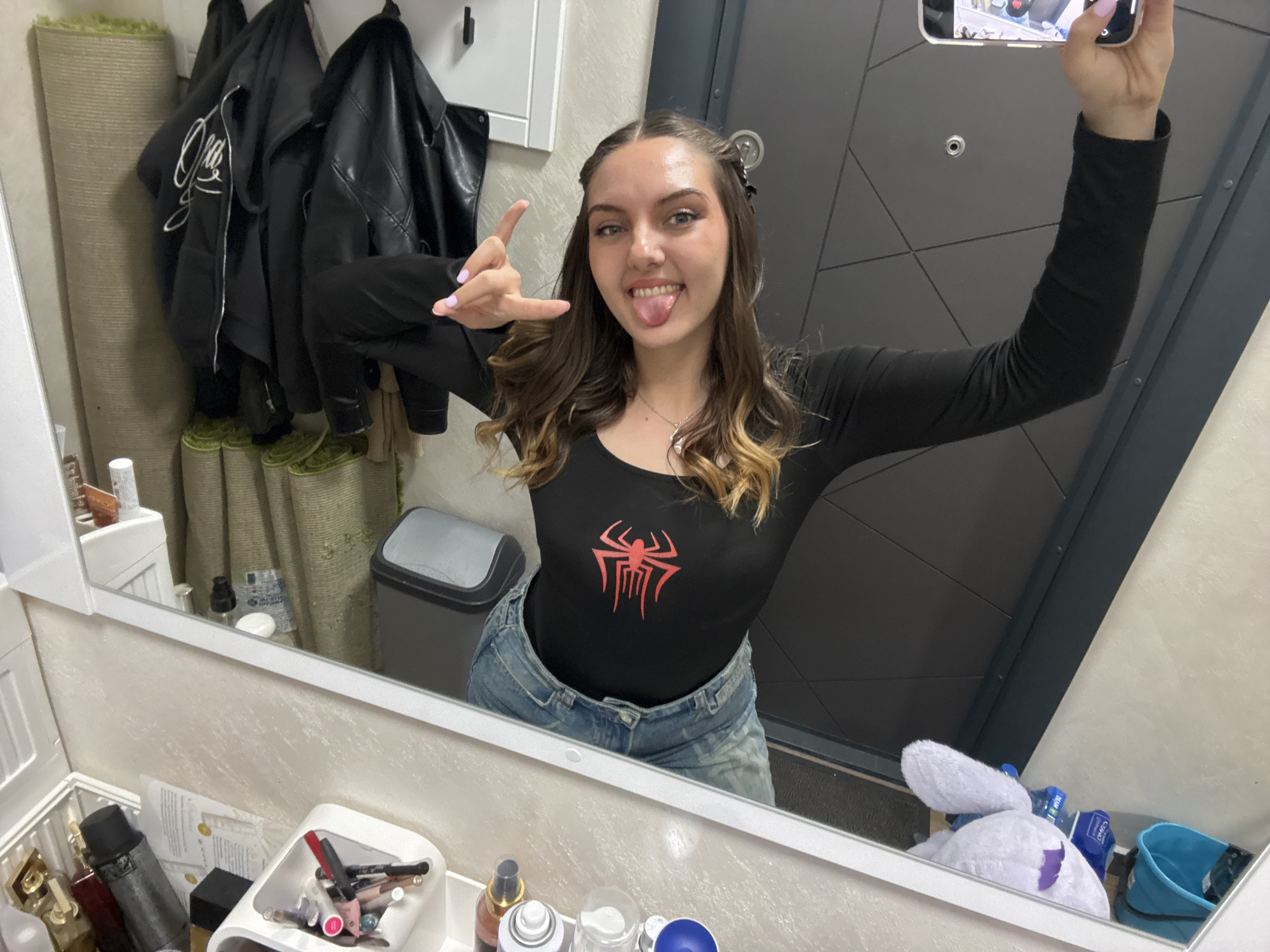 A young woman poses for a selfie in a bathroom mirror. She is wearing a long-sleeved black shirt with a red Spider-Man logo, and blue jeans. She is sticking her tongue out and making the "rock on" hand gesture. Behind her is a door with a geometric pattern, and to her left is a rack with several black leather jackets. There is a gray trash can next to the rack, and below the mirror, there are various toiletries and cosmetic items. The location appears to be inside a private residence and not discernable to Sofia, Bulgaria.