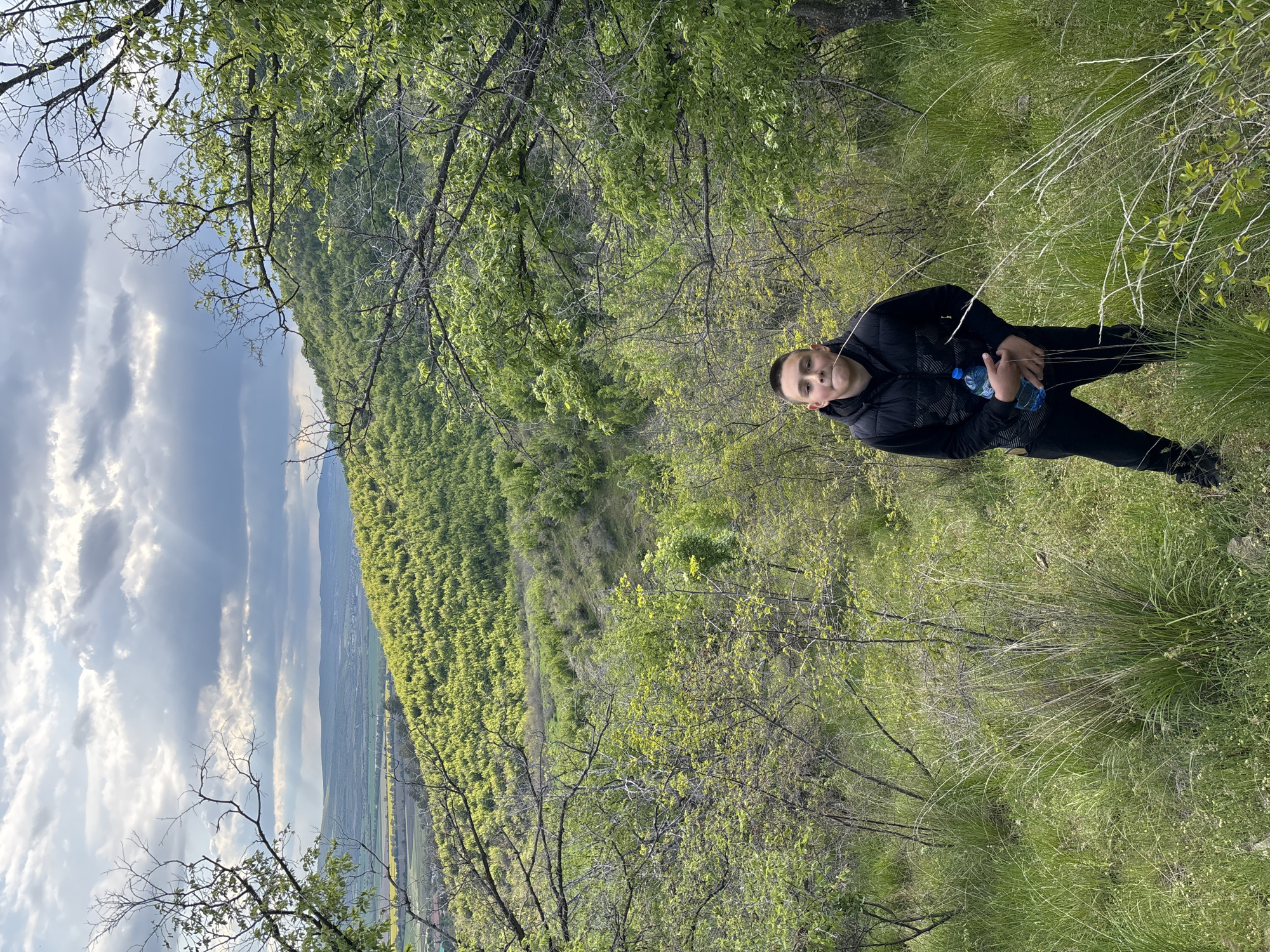 A photo shows a young person in a black jacket and pants, holding a water bottle, in a grassy, wooded area. In the background is a valley with a forest and cloudy sky. The photo was taken in Goritsa, Bulgaria.