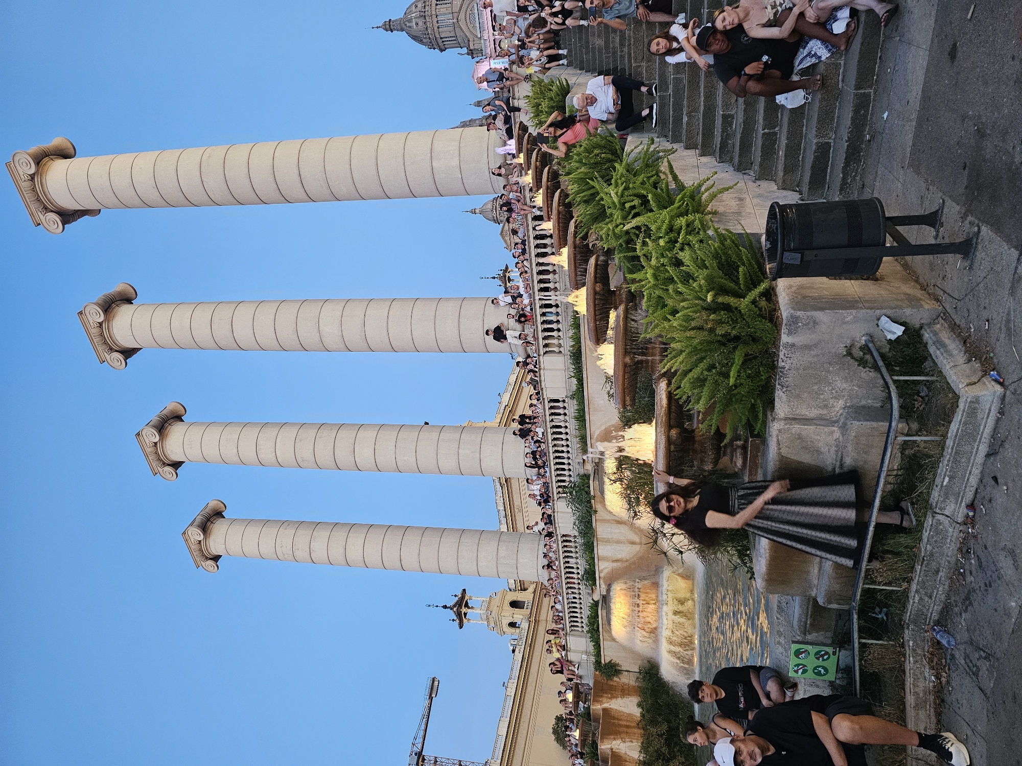 The image captures a view of the Four Catalan Columns on Avinguda de la Reina Maria Cristina in Barcelona, Spain, with people gathered around a fountain. Four large, fluted columns with Ionic capitals stand tall against a clear blue sky. In the foreground, a woman in a black dress and a metallic skirt poses near a tiered fountain, which has water flowing and illuminated jets. Several other individuals are seated on stone steps leading up to a balcony or terrace where more people are observing the scene. Lush green foliage is visible around the fountain and on the terraces. Various architectural elements of surrounding buildings, including domes and ornate facades, can be seen in the background. The ground area in the immediate foreground is paved and shows some litter.