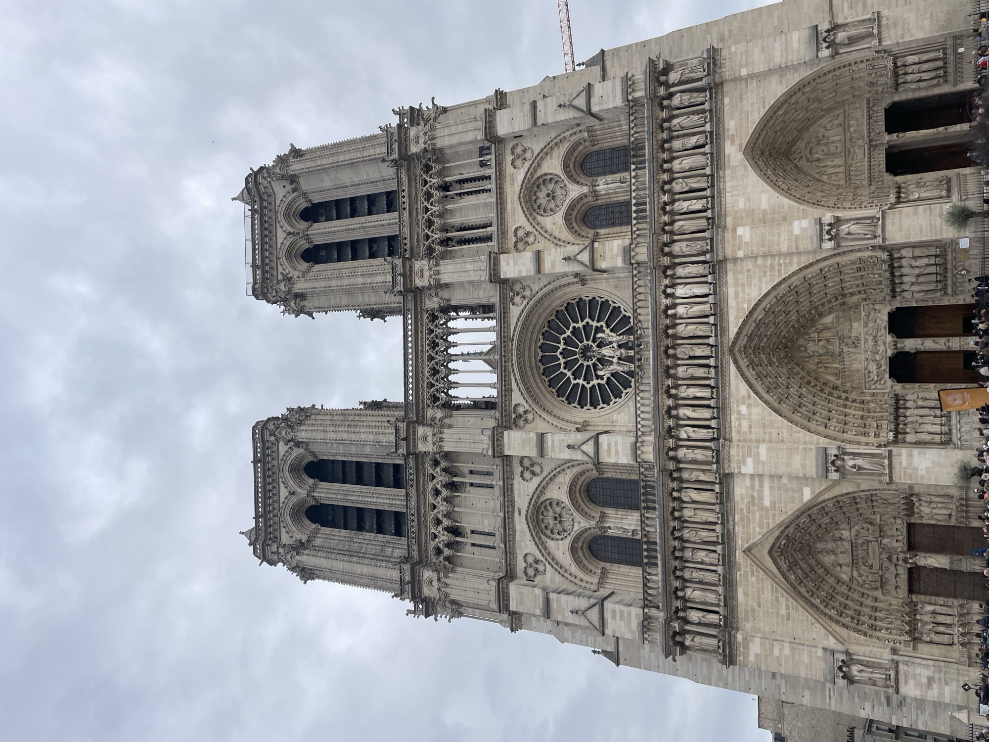 The image shows a low-angle view of the Notre Dame Cathedral in Paris, France. The facade is made of light-colored stone, with two towers visible on the left. A large rose window is centered on the facade. The main entrance, located on the right, is adorned with arches and sculptures. A crane is visible above the cathedral. A crowd of people is on the right of the image. The sky is overcast.