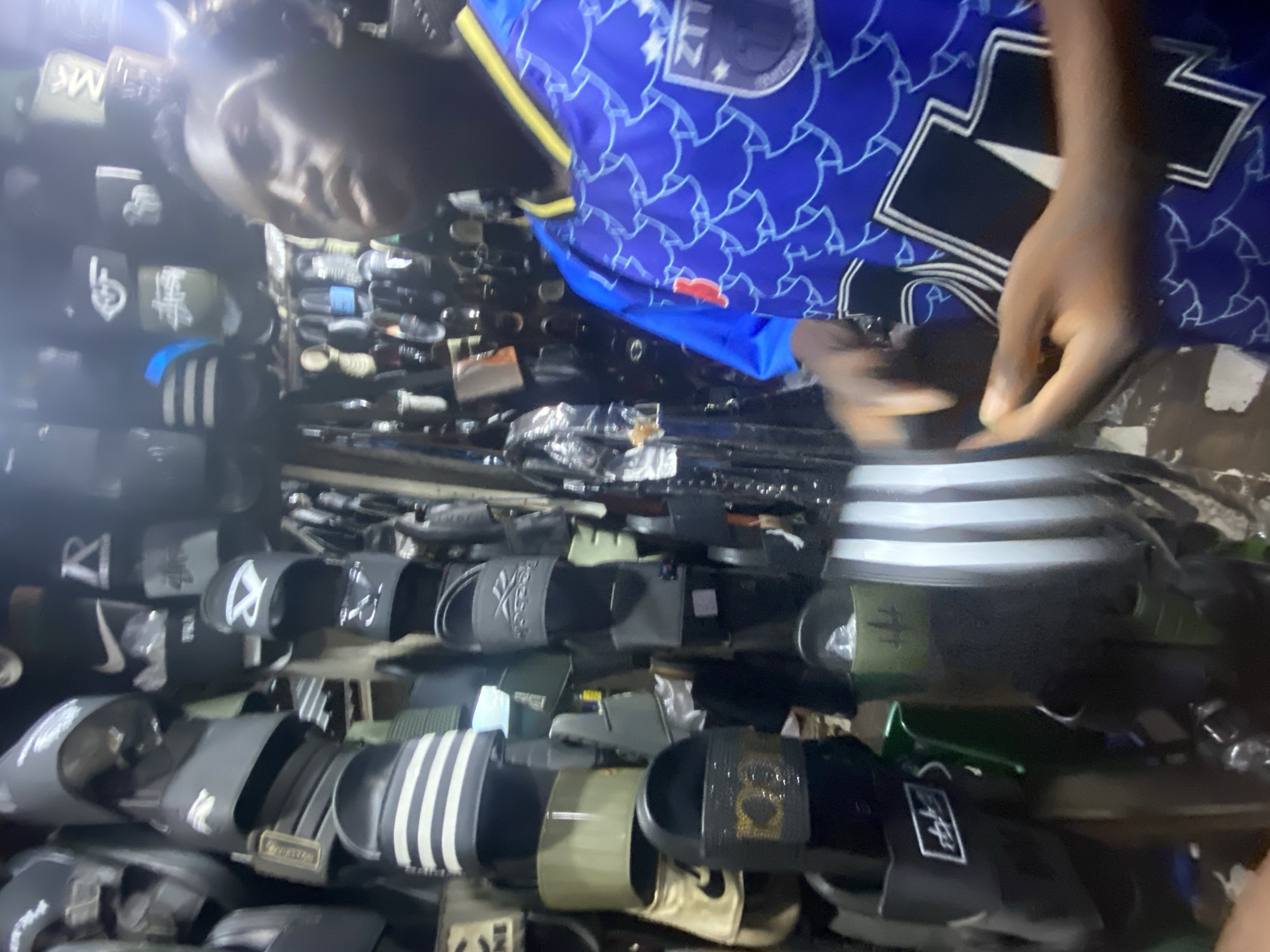 The image appears to be taken indoors, possibly at a market stall or small shop in Lagos, Nigeria. A person wearing a blue soccer jersey is interacting with a stack of various slides/sandals. The footwear is of different brands, including Adidas, Nike, and Reebok, among others. The slides are stacked high in the foreground, with rows of additional footwear extending into the background. Other merchandise is also visible, obscuring the wall behind. The lighting is dim, typical of a small retail space. The person’s hands are the only body parts in focus, examining the slides.