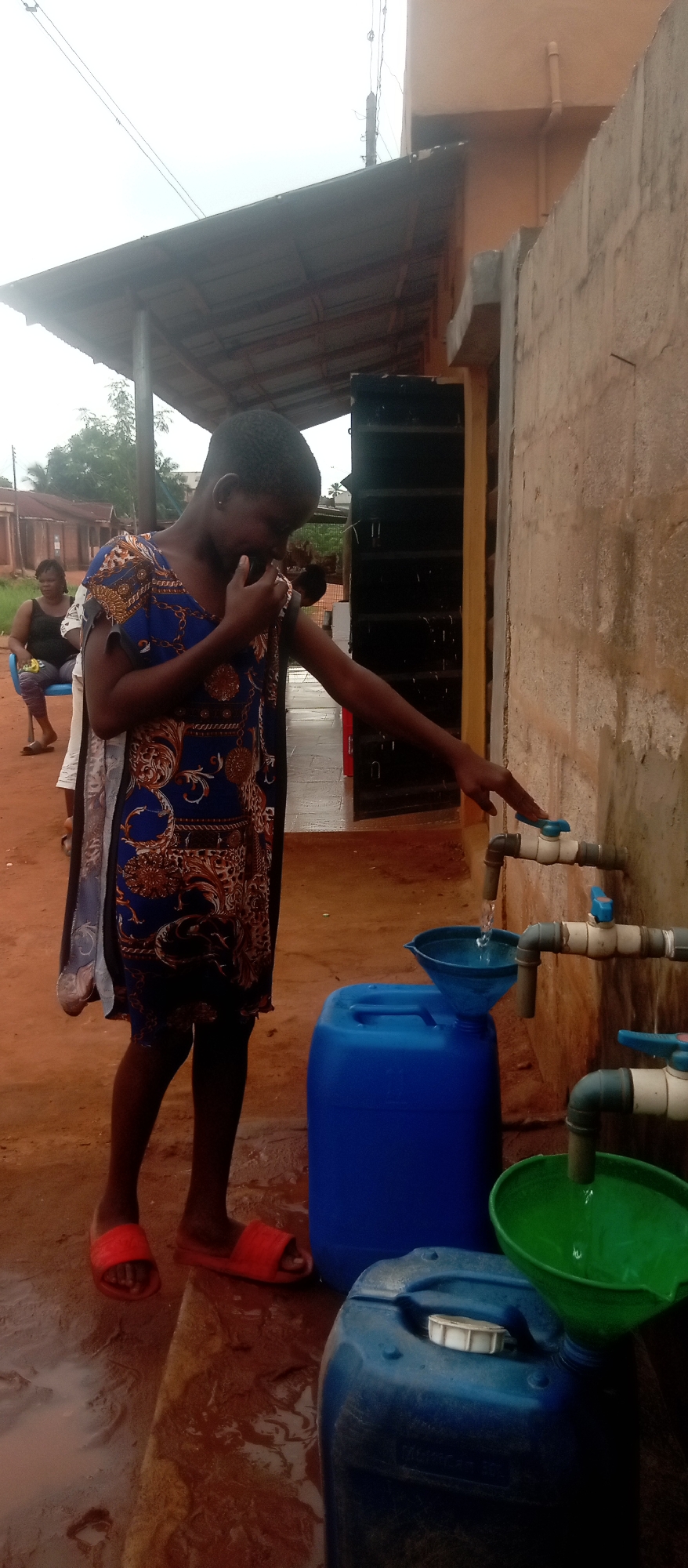 The image shows a young girl in Ogidi, Nigeria, standing in front of a wall with three faucets. She is touching the handle of the top faucet, from which water is flowing into a blue funnel placed over a blue container. Another faucet is directing water into a green funnel over another blue container. A woman is sitting on a blue chair in the background. The ground is wet and appears to be made of red dirt or clay. The wall is rough and textured. The lighting suggests an overcast day.