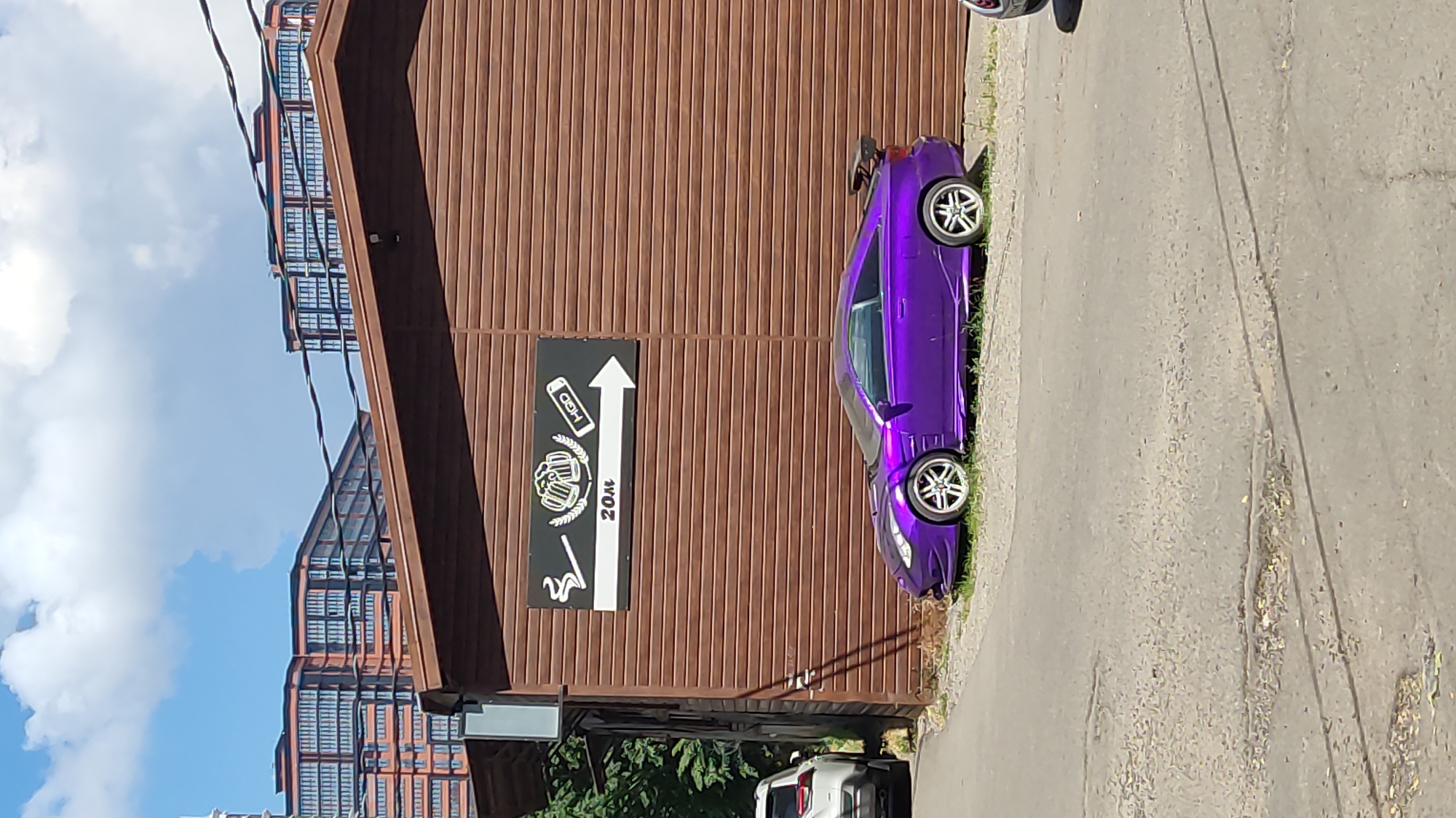 A vibrant purple sports car is parked on a patch of gravel next to a long, brown, corrugated building. Above the car, on the building's wall, is a rectangular sign with white and black graphics. The sign depicts a cigarette, a beer mug, and a tube with the letters "HGD". Below these graphics, the number "20m" is visible, followed by a white arrow pointing to the right. In the background, a white SUV is partially visible on the left, and in the far background, several multi-story apartment buildings with numerous windows are visible under a blue sky with scattered clouds. The car appears to be a Toyota Celica. The scene is likely in the city of Krasnodar, Russia, given the context of the provided prompt, although this cannot be visually confirmed from the image itself.