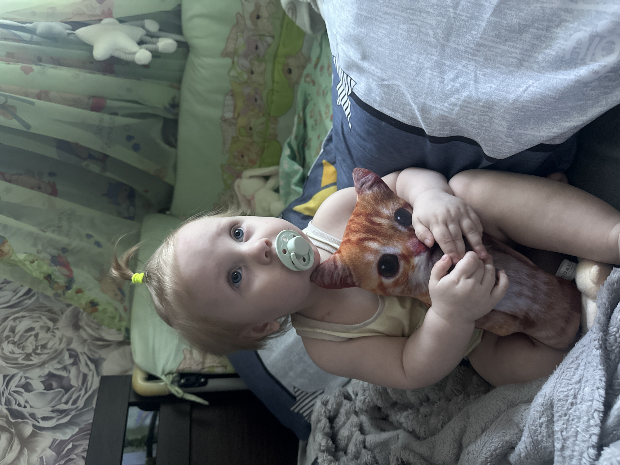 A young child with blond hair pulled into a small ponytail and blue eyes is sitting on a bed, clutching a stuffed cat toy. The child has a light blue pacifier in their mouth. The child is wearing a yellow tank top. The bed has dark blue and gray patterned bedding. In the background, there are sheer green curtains with a pattern of animals, and a white stuffed star hanging. A floral wallpaper is visible on the wall behind the bed. The overall environment appears to be a child's bedroom. The location is identified as Sayanogorsk, Russia.