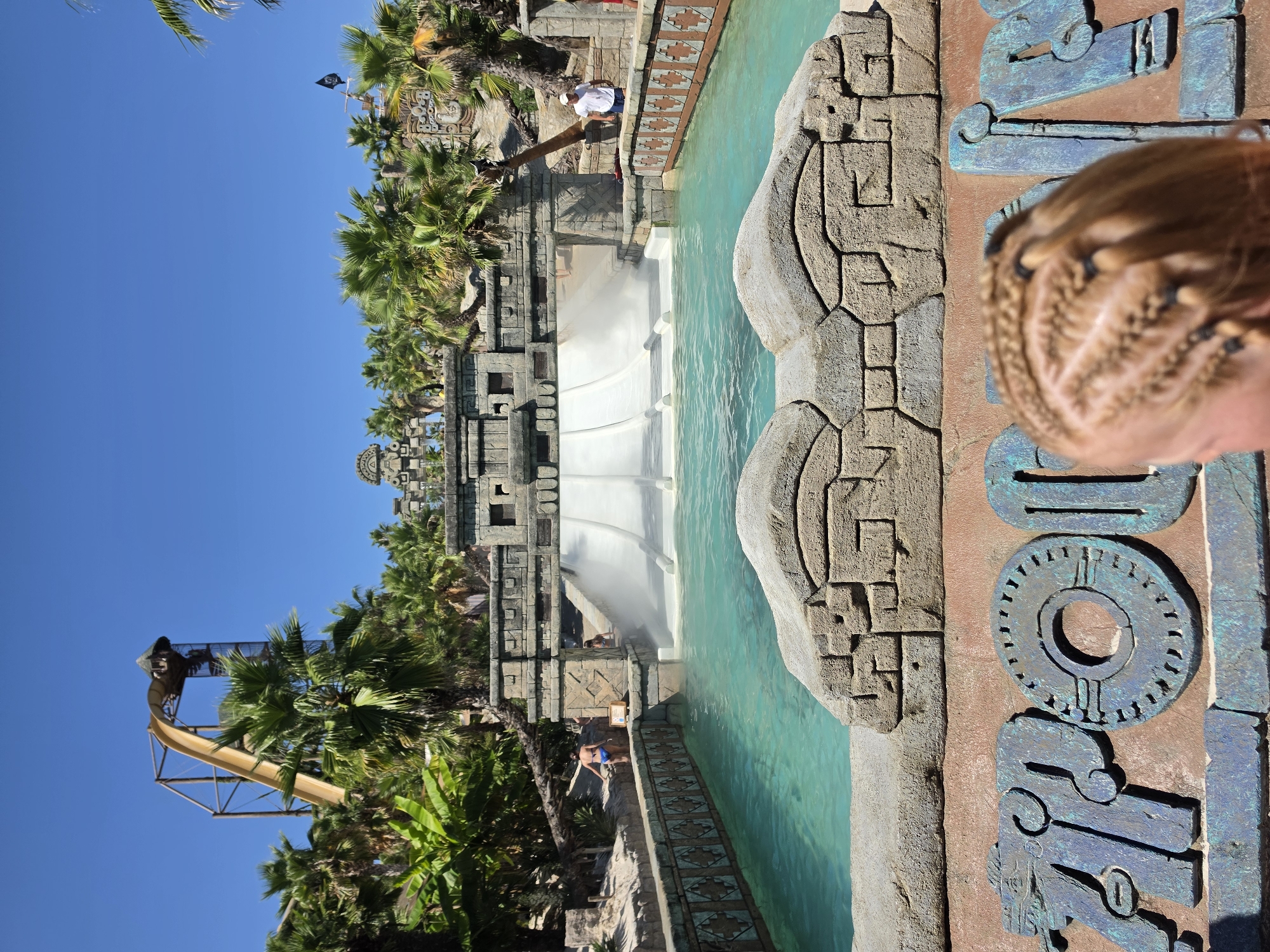 The image captures a sunny day at Aqualandia water park in Jesolo, Italy, showcasing a thrilling water slide complex with an Aztec theme. In the foreground, the upper part of a person's head, with braided blonde hair, is visible. In the mid-ground, a wide water slide with multiple lanes descends into a turquoise pool. The slide structure is made of stone, decorated with intricate carvings, resembling ancient ruins. Palm trees and lush vegetation surround the area, enhancing the tropical atmosphere. In the background, a tall, yellow water slide stands against a clear blue sky. People are seen enjoying the water park, with some walking by the pool and others preparing to use the slides. The overall scene depicts a lively and enjoyable day at a water amusement park.